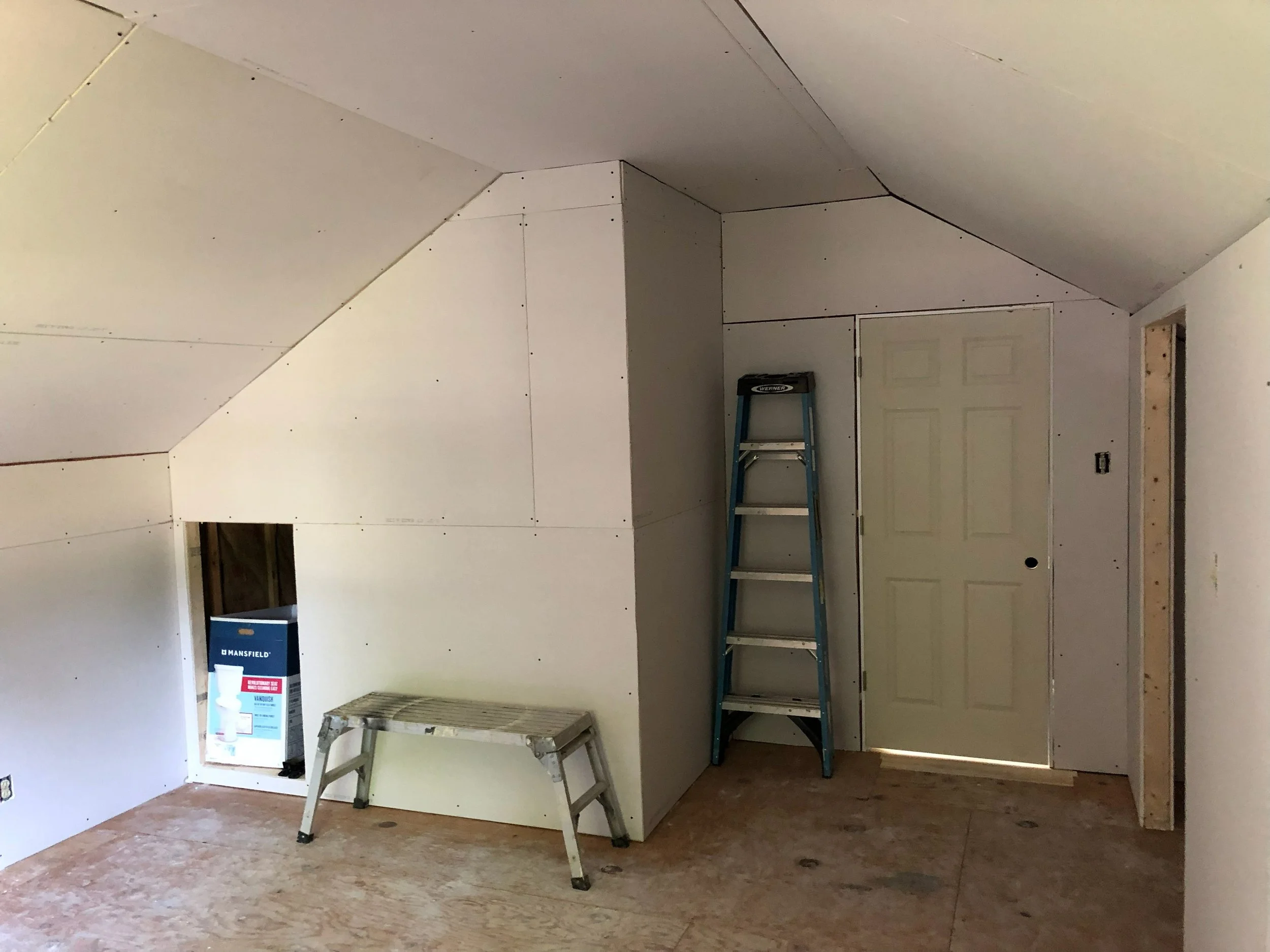 Interior of a room under construction with drywall installed on the walls and ceiling, a small stepladder, a blue step ladder near a door, and an open window with a box inside.
