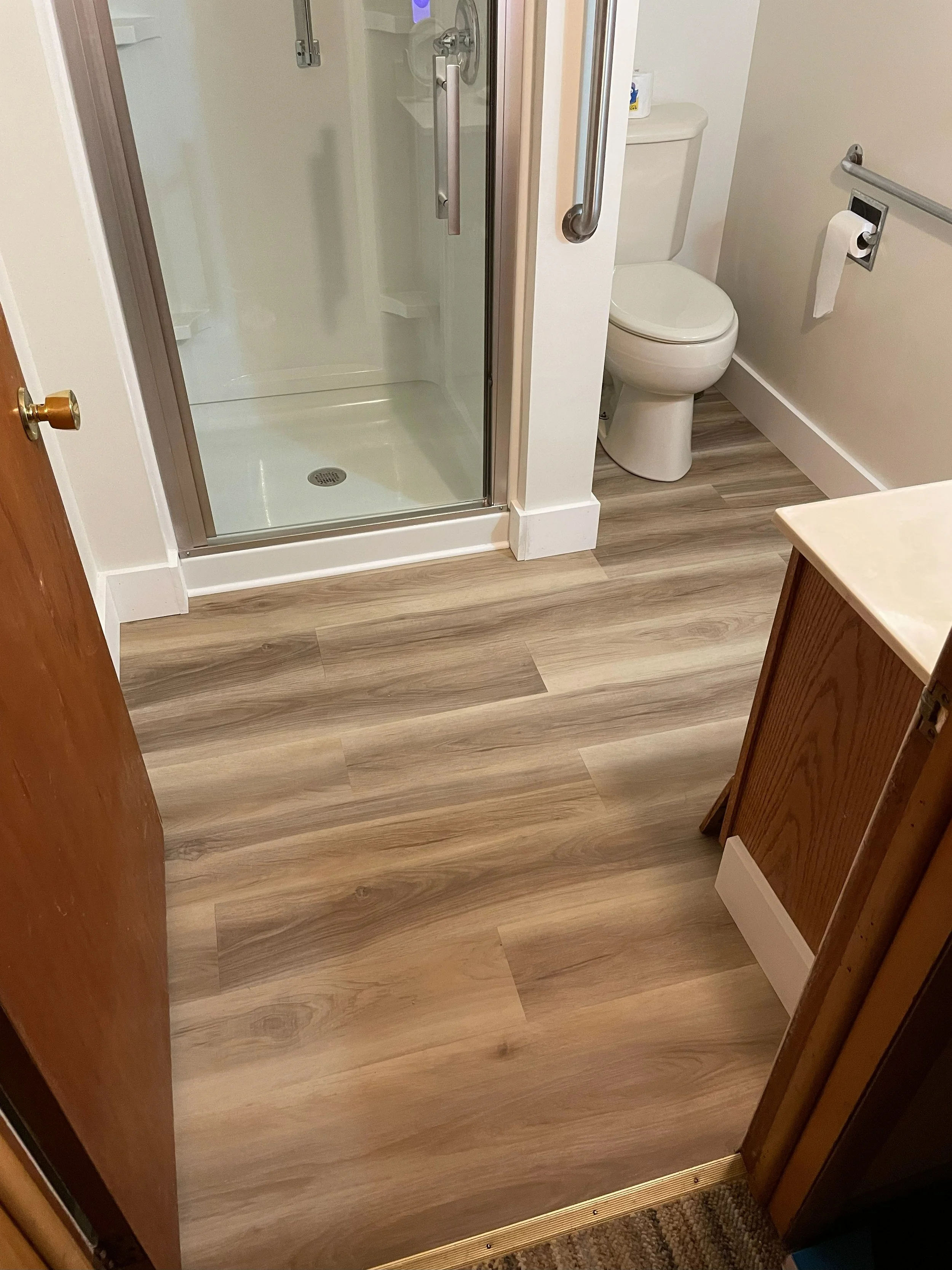 View of a bathroom with a wooden door open, showing a shower with a glass door, a toilet with a roll of toilet paper, and a wood cabinet on the right side.