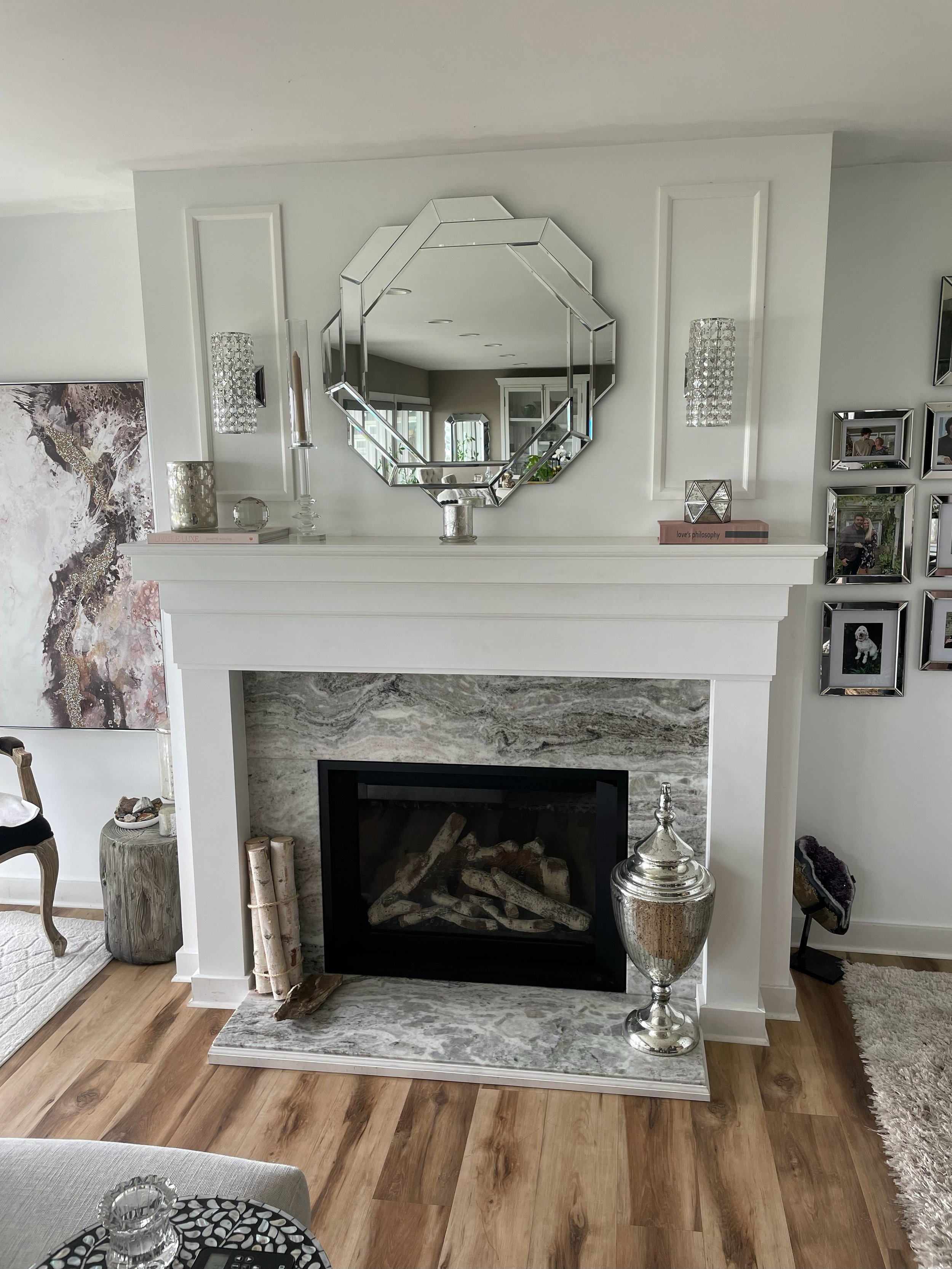 Living room fireplace with a white mantel and marble surround, topped by a large octagonal mirror, with decorative items and family photos on the mantel.