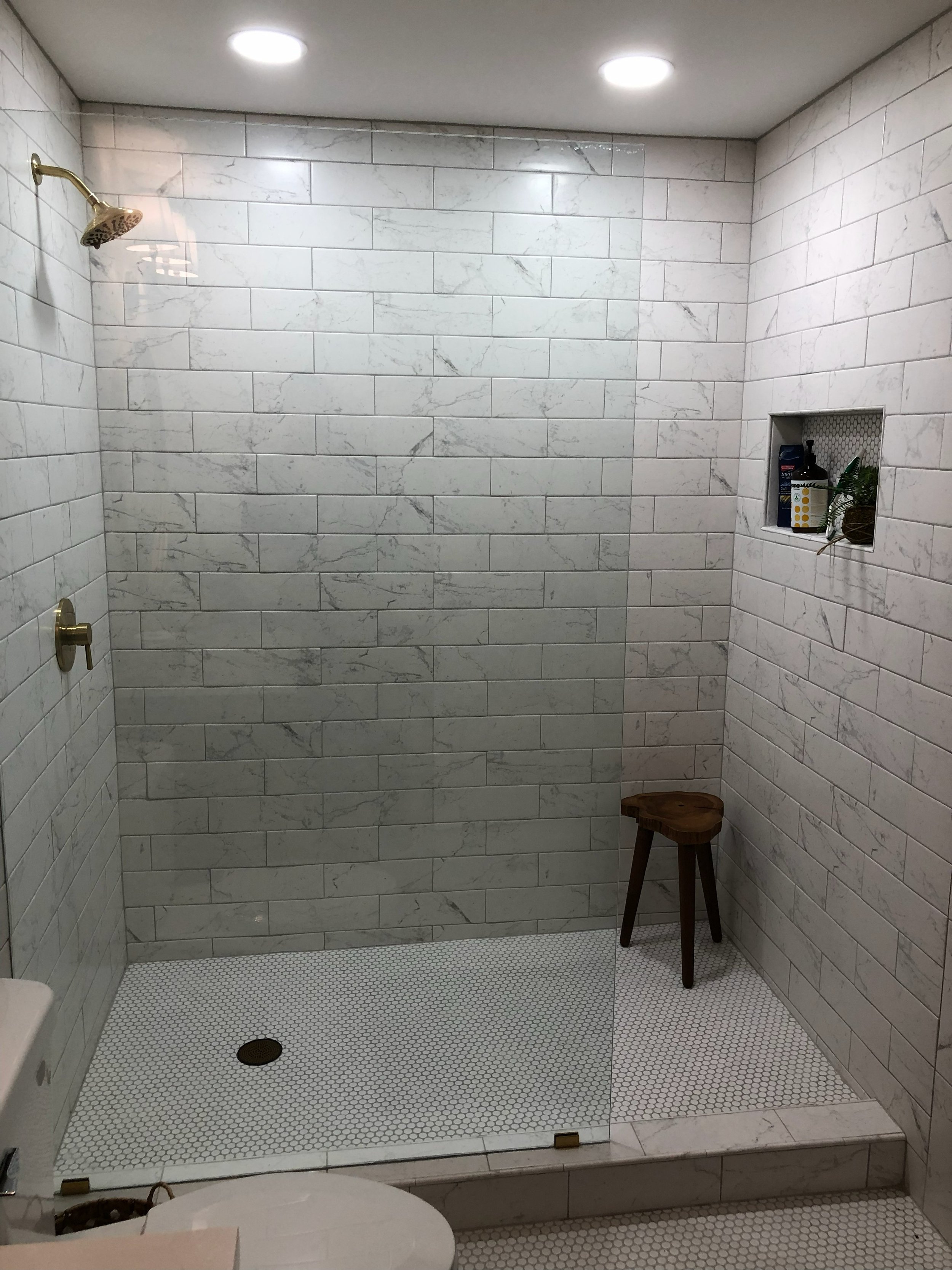 A minimalist walk-in shower with white marble tile walls, two gold shower fixtures, a small nook with toiletries, a wooden stool, and lighting from two ceiling lights.