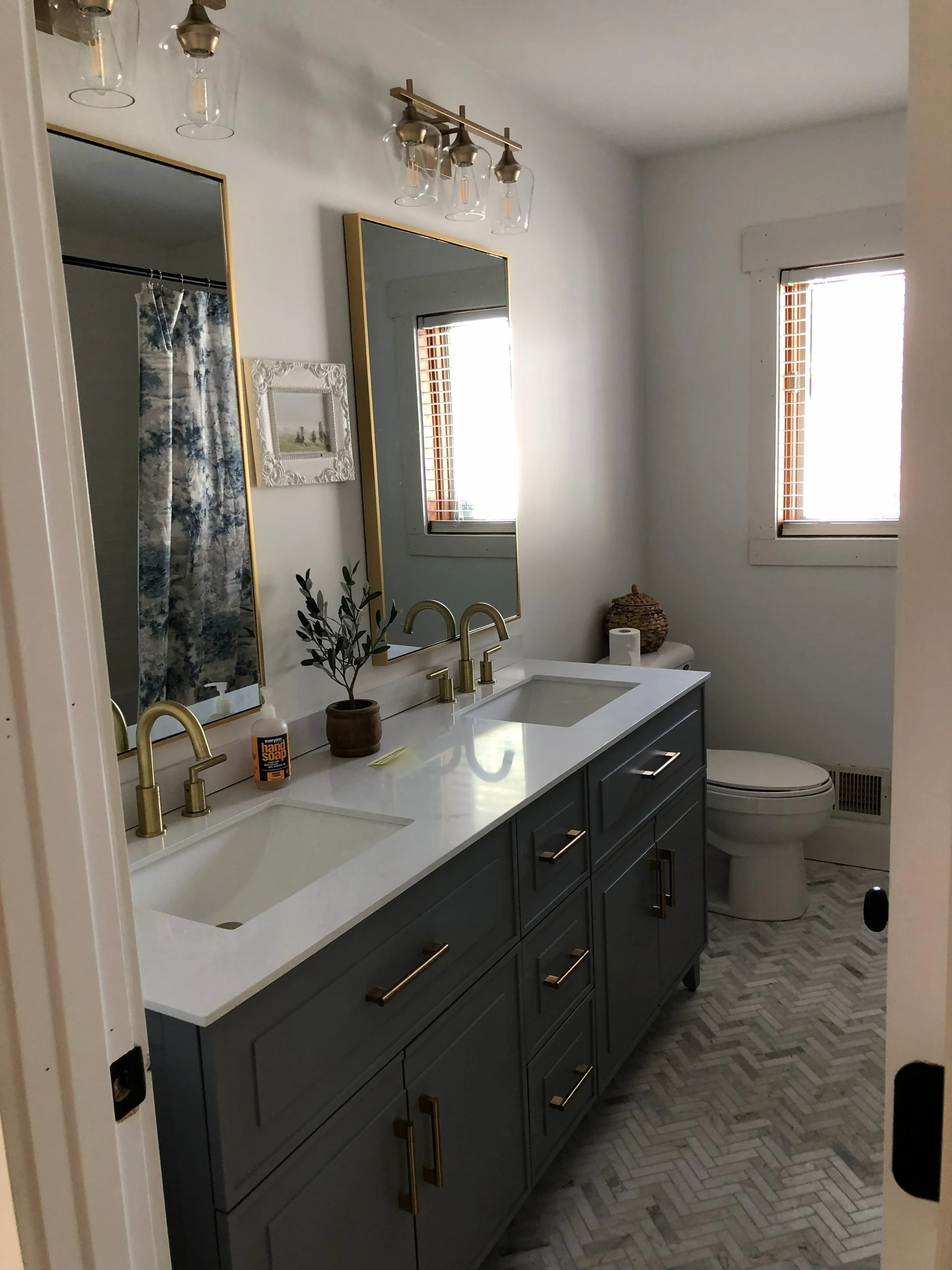 Modern bathroom with dual sinks, gray vanity, gold fixtures, mirrors, small decorative plants, windows with blinds, and a toilet.