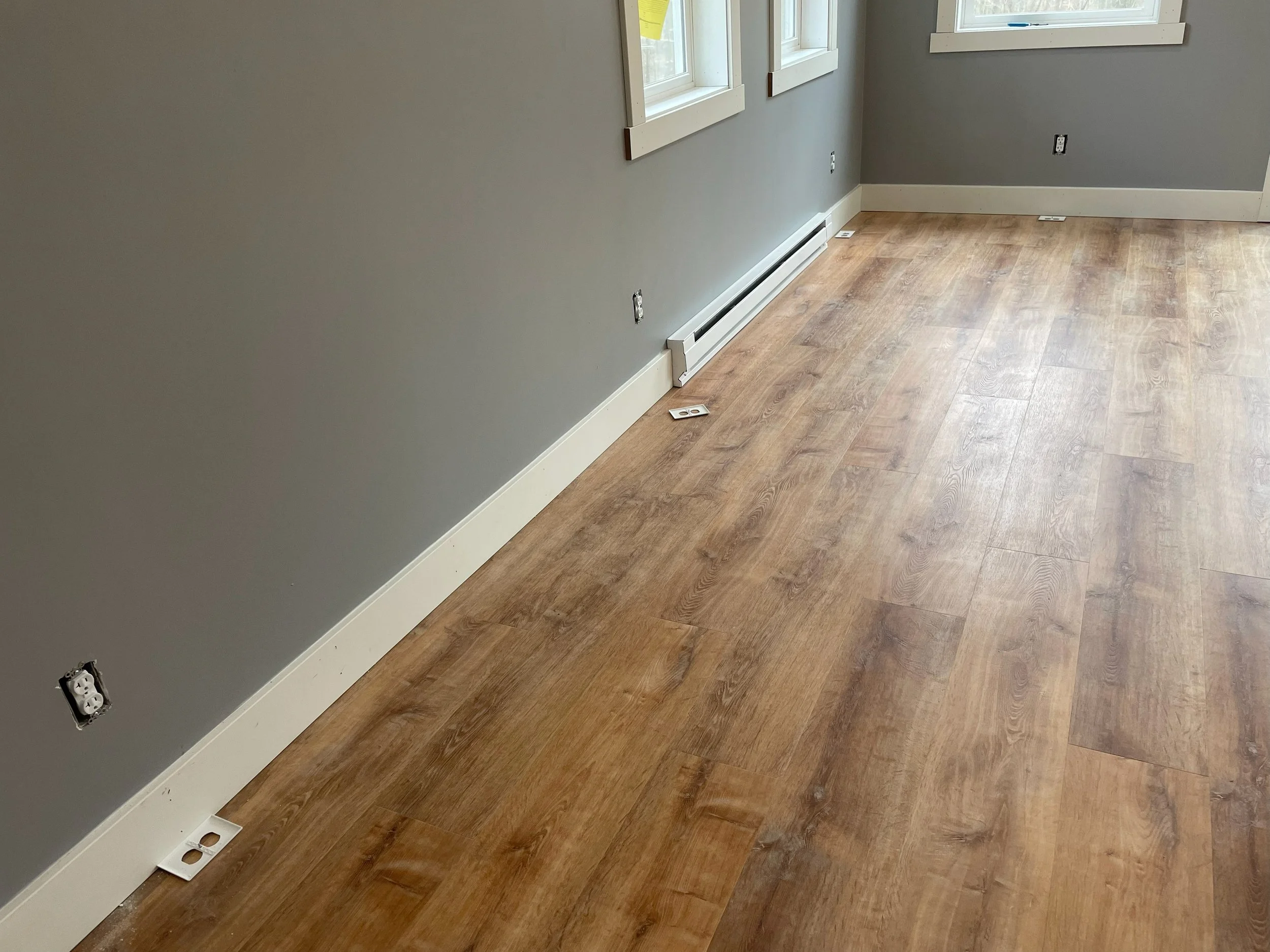 Empty room with gray walls, hardwood flooring, electrical outlets, and baseboard heater. Three small white electrical boxes are on the floor near the wall.