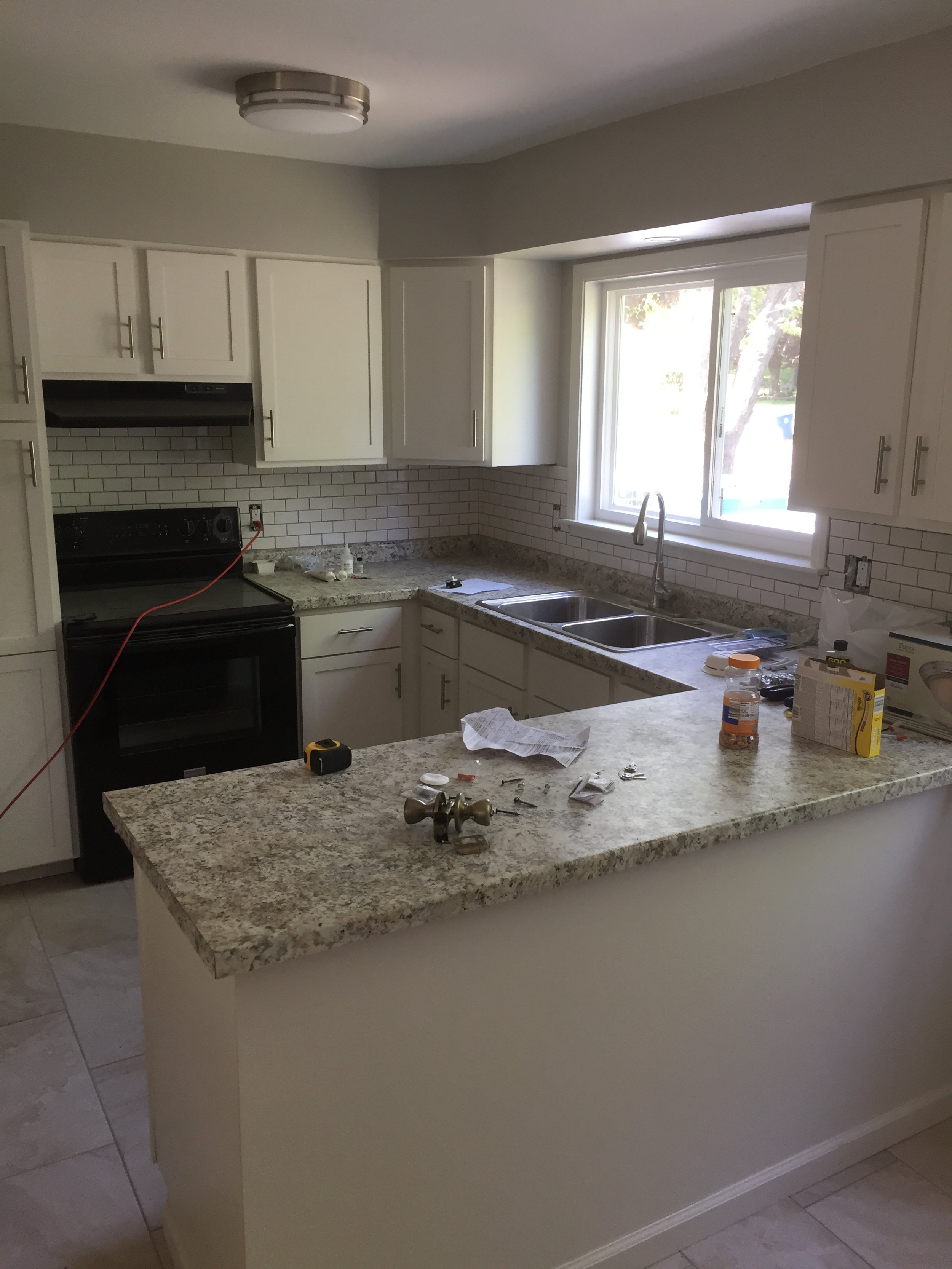 Kitchen under renovation with tools, paper, and supplies on granite countertop, white cabinets, double sink, window, and black stove.