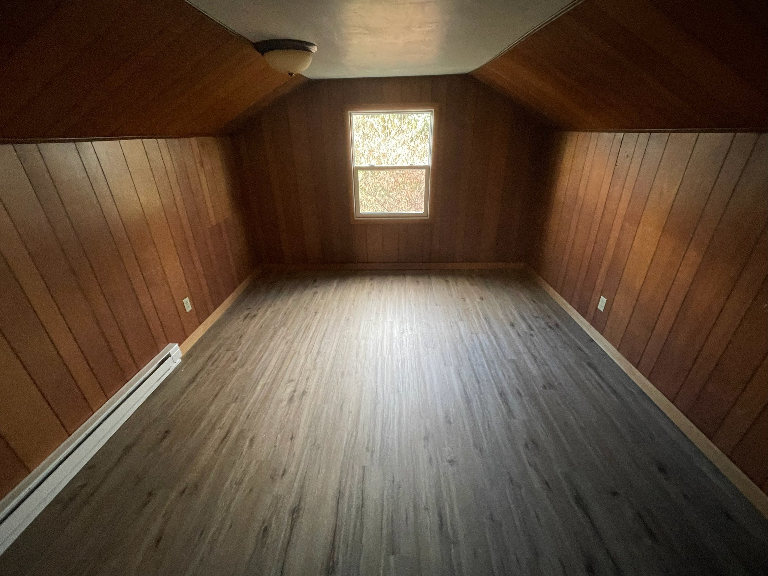 Empty room with wood-paneled walls, a single window, and a wood floor.