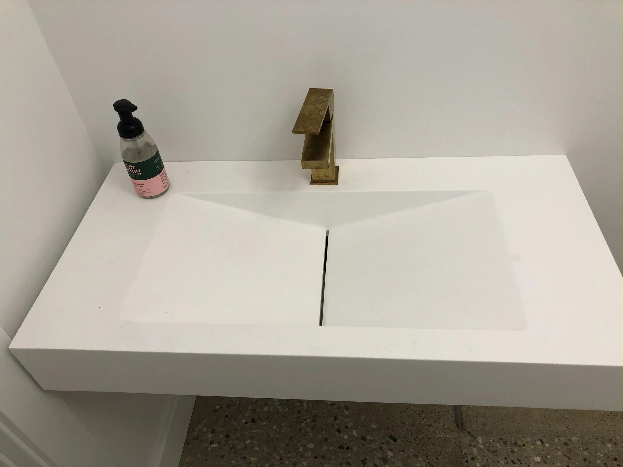 A white bathroom sink with a gold-colored faucet, a bottle of liquid soap on the left, and a small golden tray above the sink.