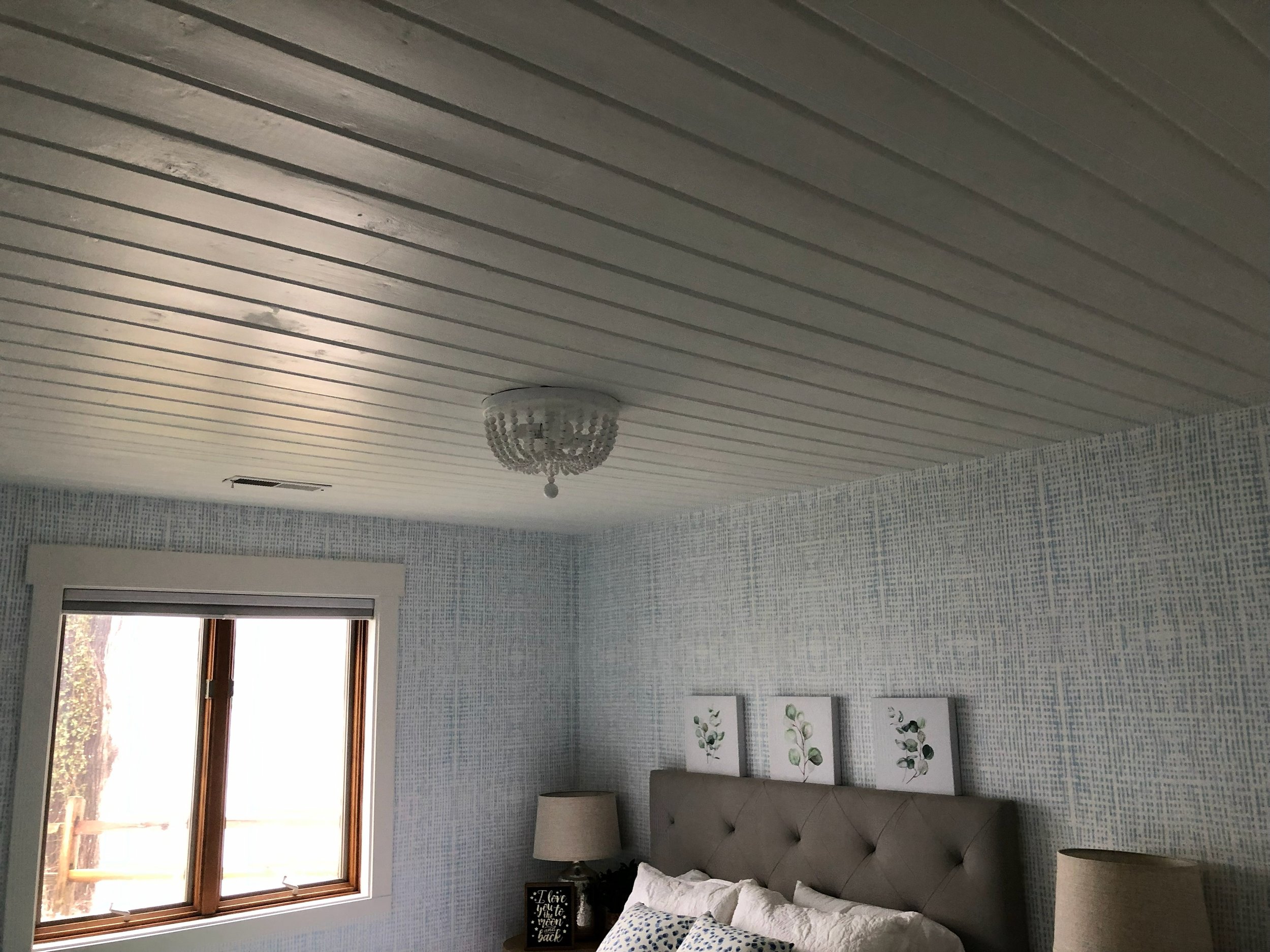 A bedroom with a window on the left, a beige tufted headboard, white bedding, two nightstands with beige lamps, and three framed botanical prints on the wall. The ceiling has light-colored wooden planks and a decorative chandelier.