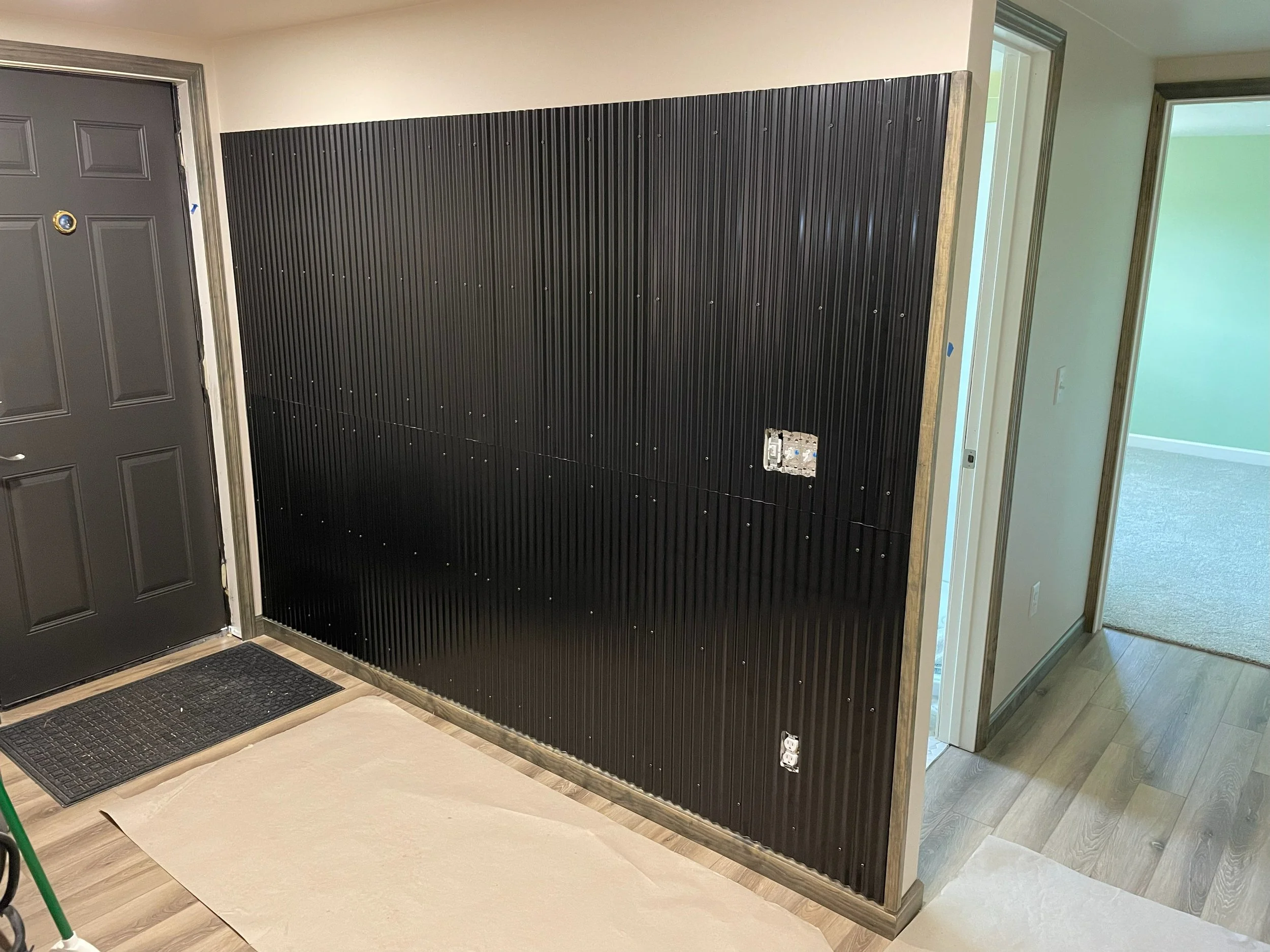 Interior of a home with a black wall panel and electrical outlets, next to a door and adjacent room with green walls and carpet.