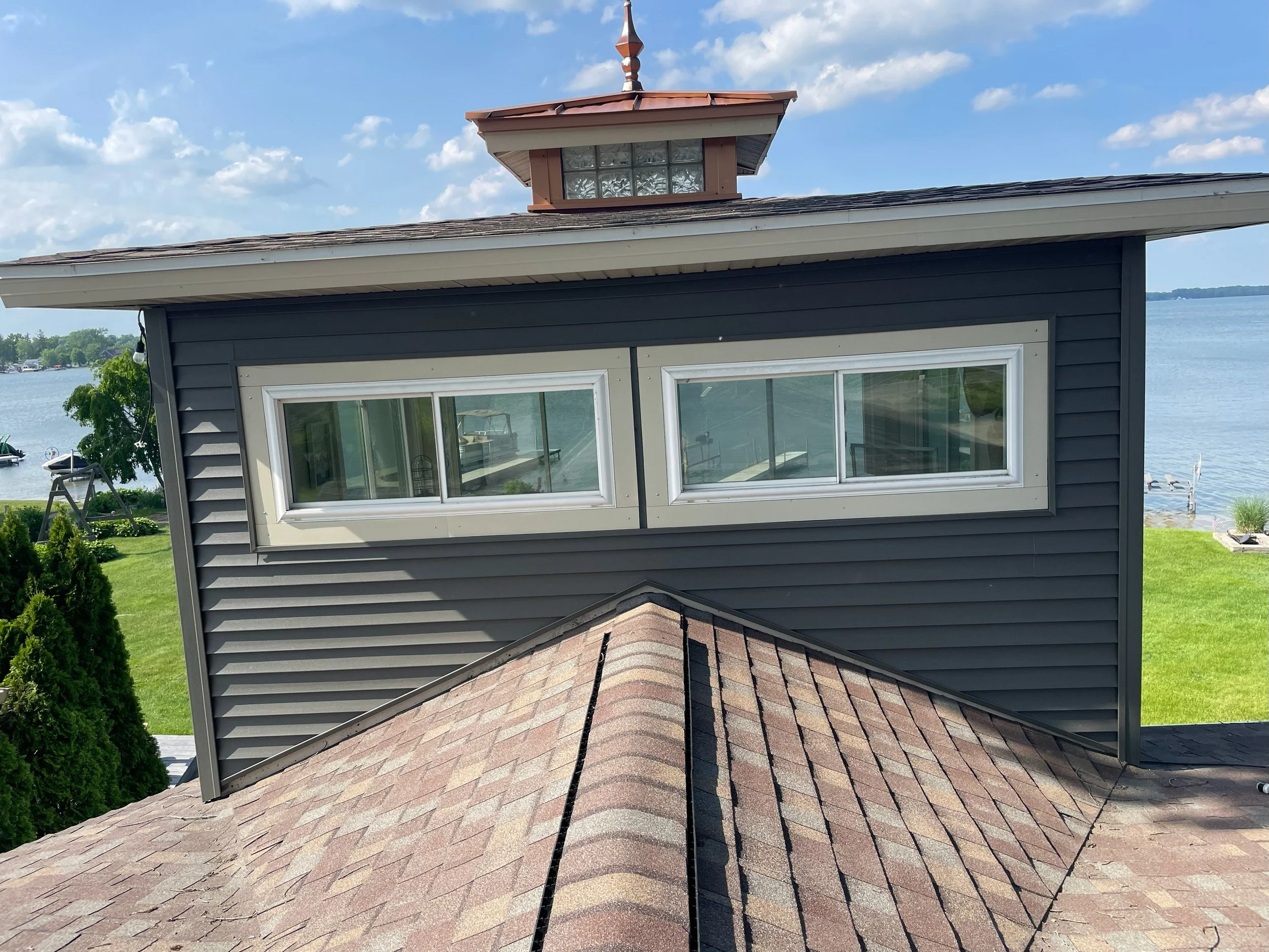 View of a dark grey house with a sloped asphalt shingle roof, a small cupola on top, and a large horizontal window. There is a lake and a grassy yard with trees and a boat dock in the background.