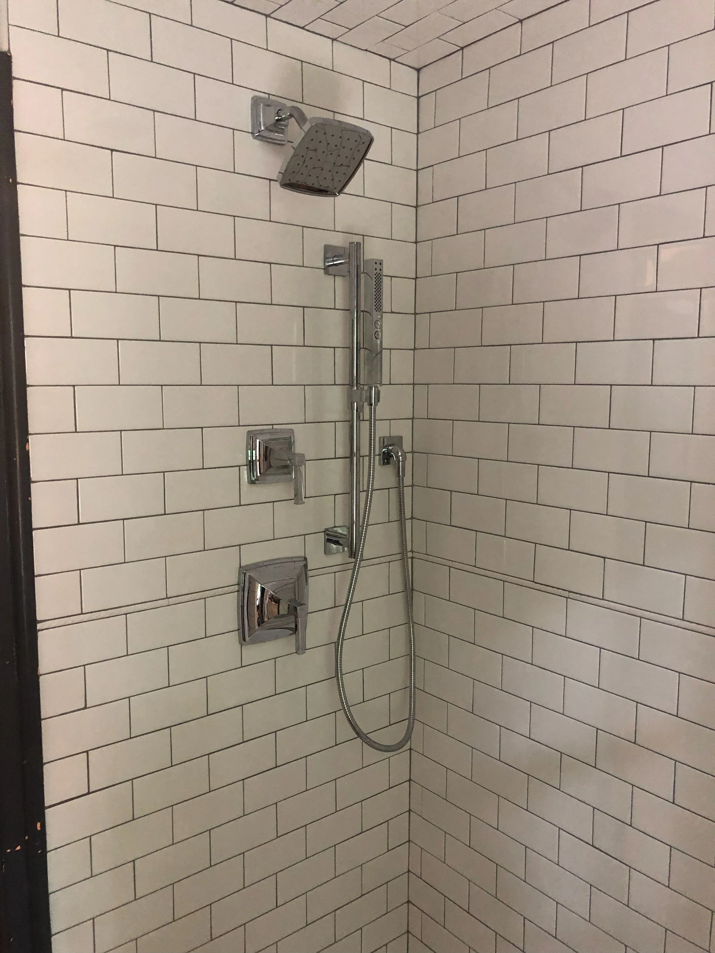 Shower area with beige subway tiles, chrome fixtures, a rainfall showerhead, a handheld shower, and three control handles.