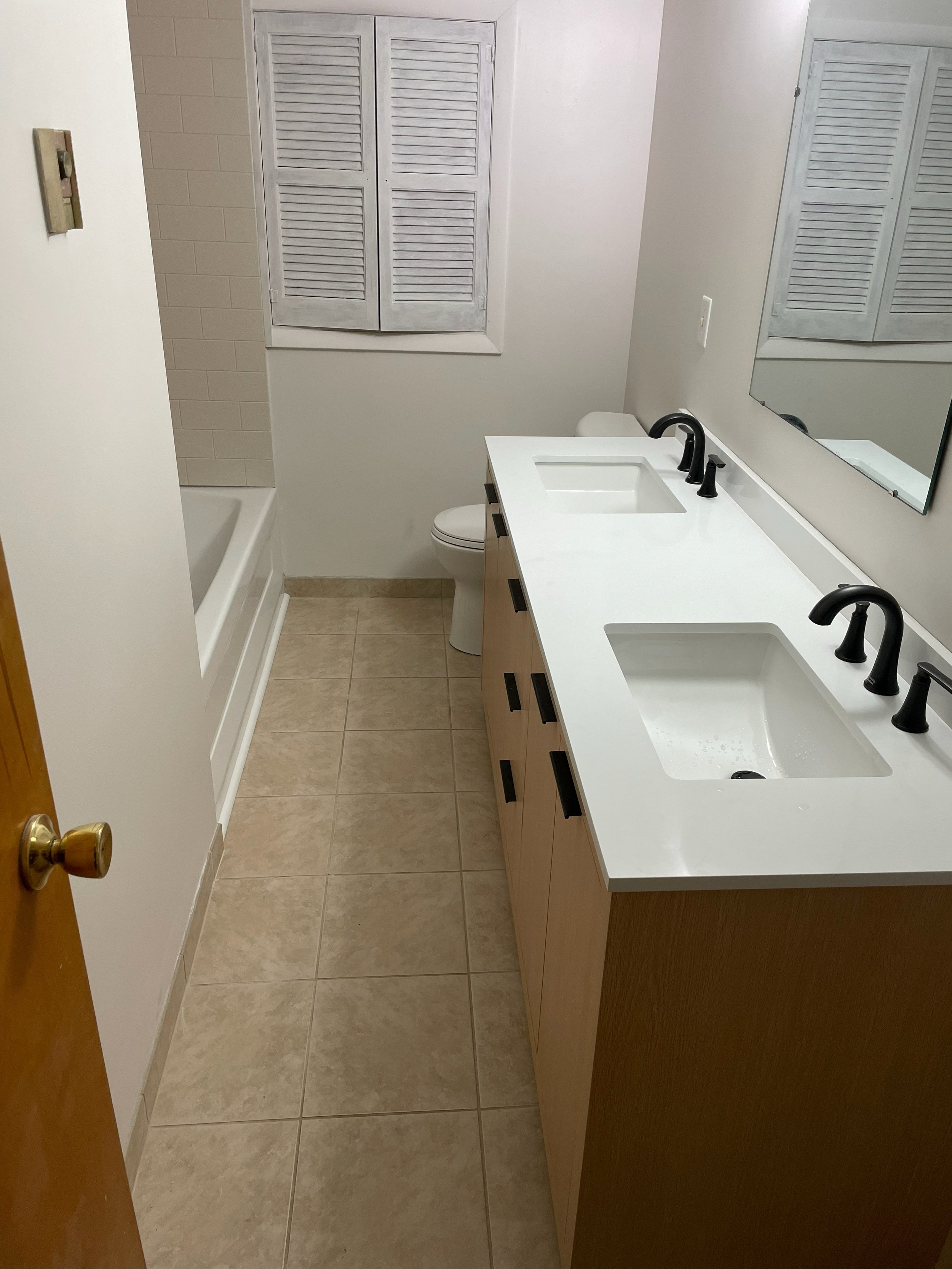 A bathroom with a double sink vanity, black faucets, large mirror, toilet, bathtub, and a window with closed blinds.