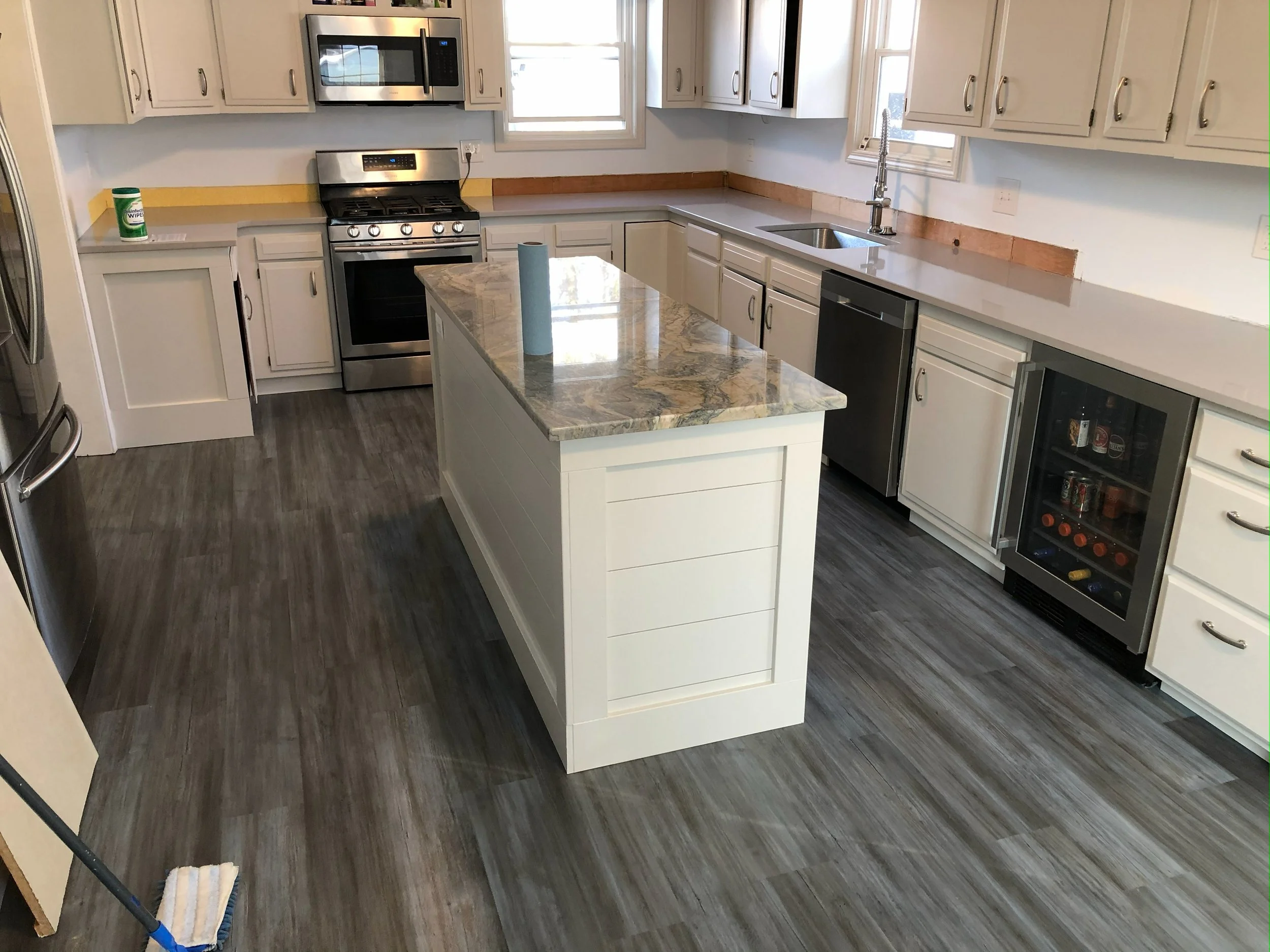 Kitchen with white cabinets, stainless steel appliances, a marble-topped island, gray wood-look flooring, and a window above the sink