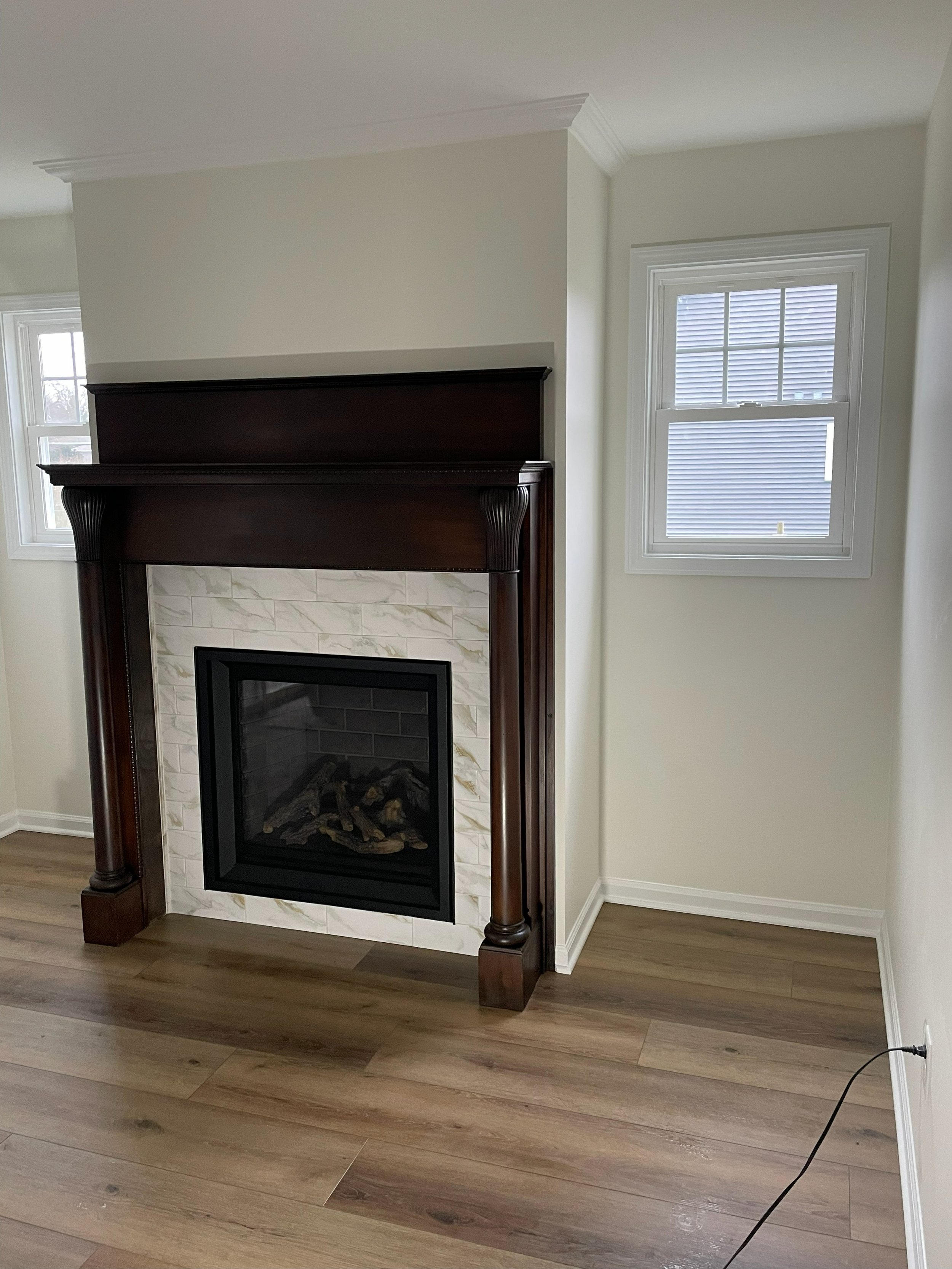 Living room corner with a fireplace framed by dark wood mantel and white marble tiles, two windows on adjacent walls with white trim, wood flooring, and a black electrical cord on the floor.