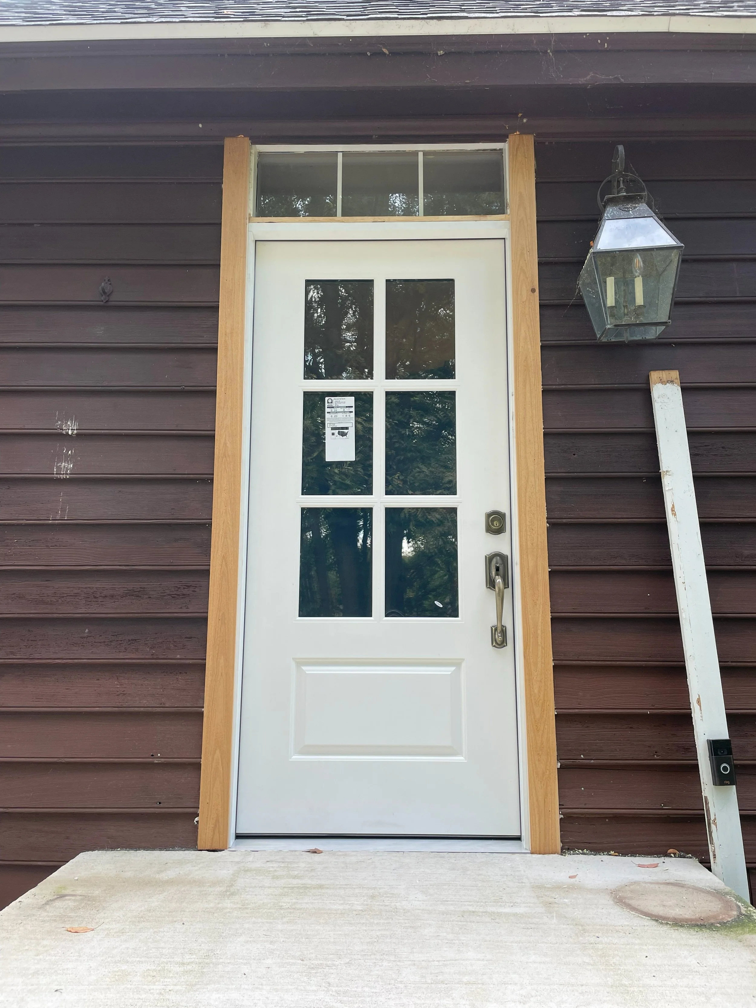 White front door with window panes, brown wooden trim, dark brown siding, a lantern-style outdoor light fixture, and a step leading to the entrance.