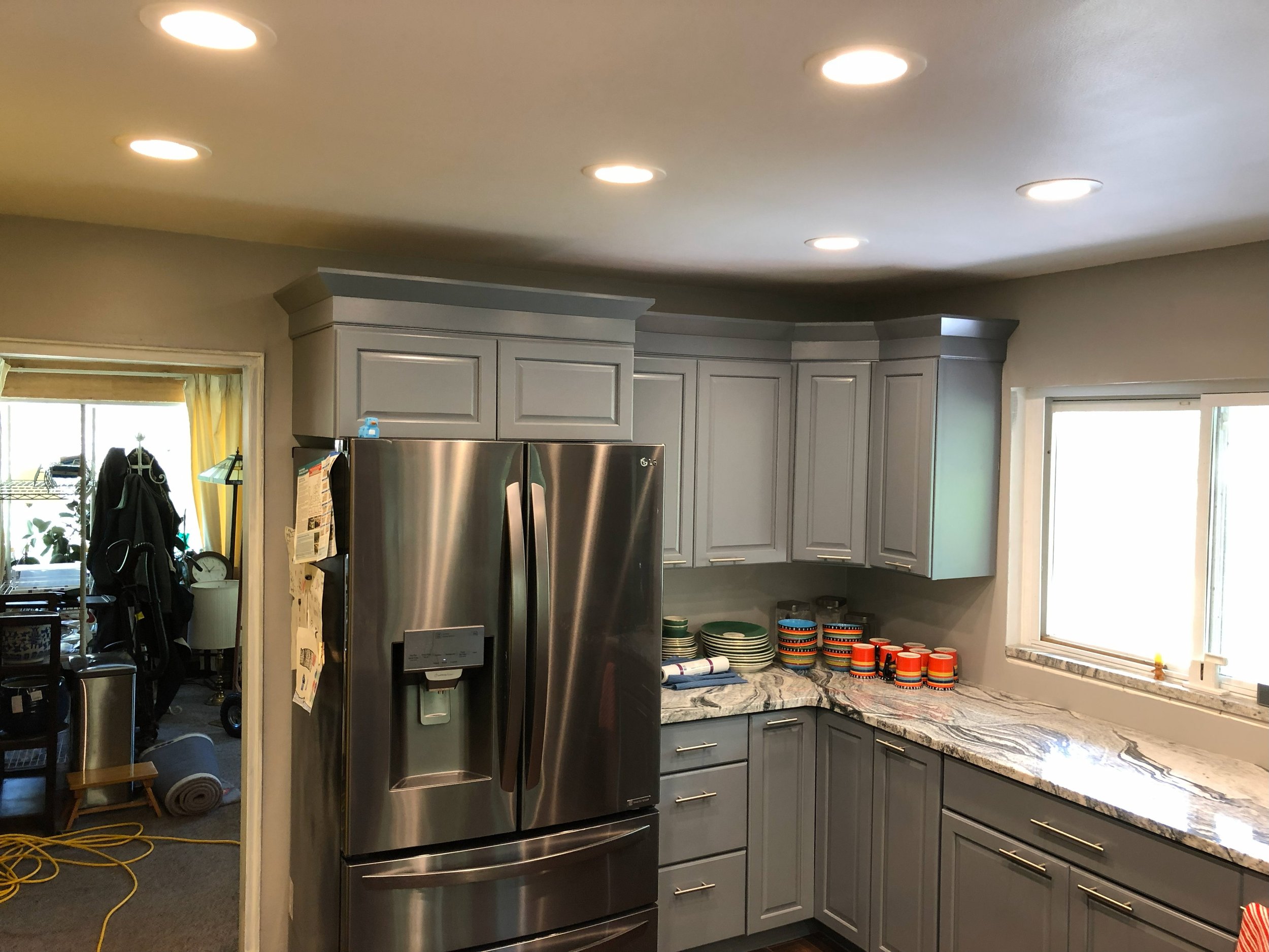 Kitchen with gray cabinets, marble countertop, stainless steel refrigerator, and window with sunlight, colorful canisters on countertop, and adjacent room with coats and items.