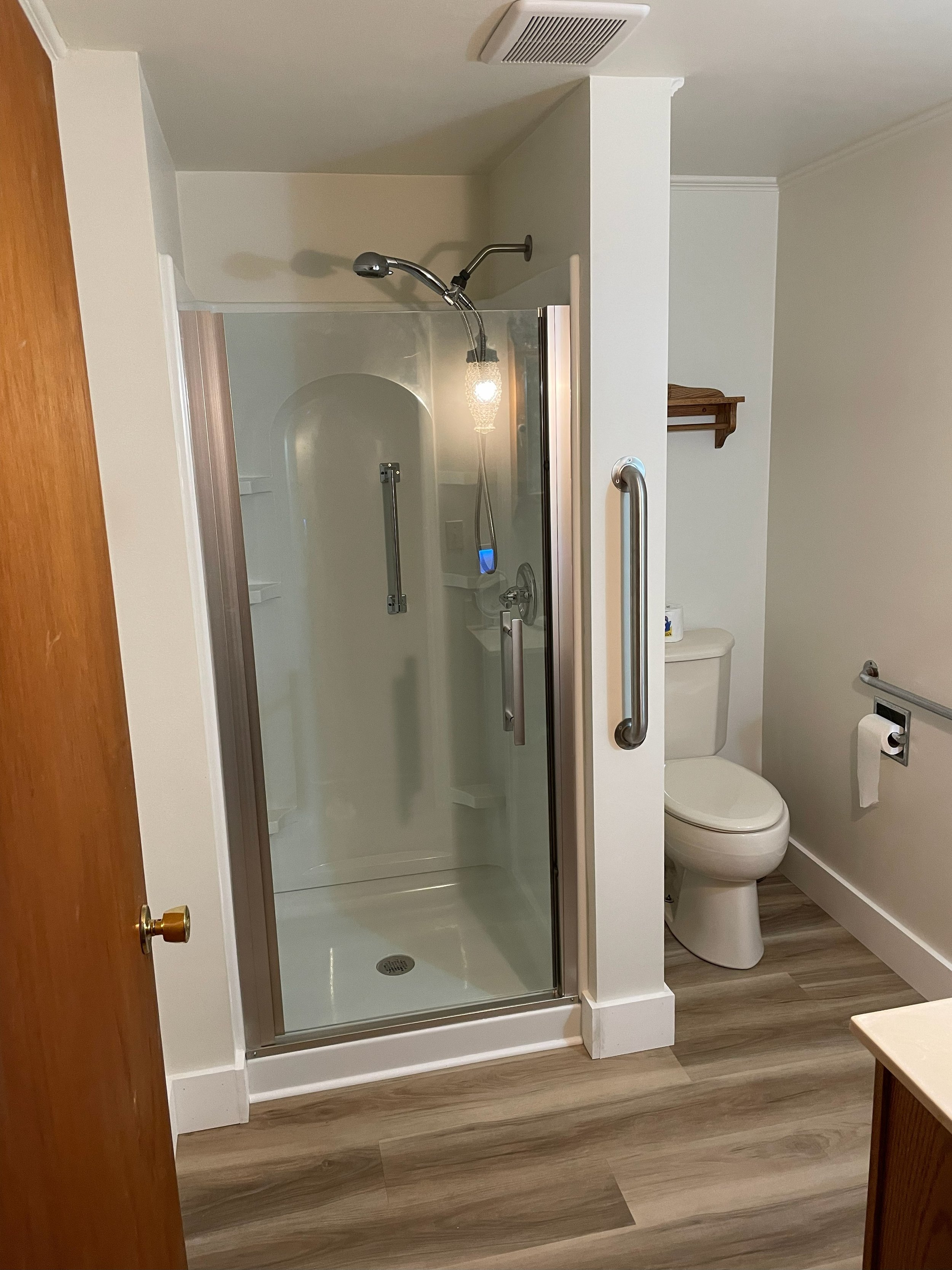 Bathroom with a glass shower stall that has a chair lift on one side, a toilet with a roll of toilet paper, wooden flooring, and a small wall shelf.