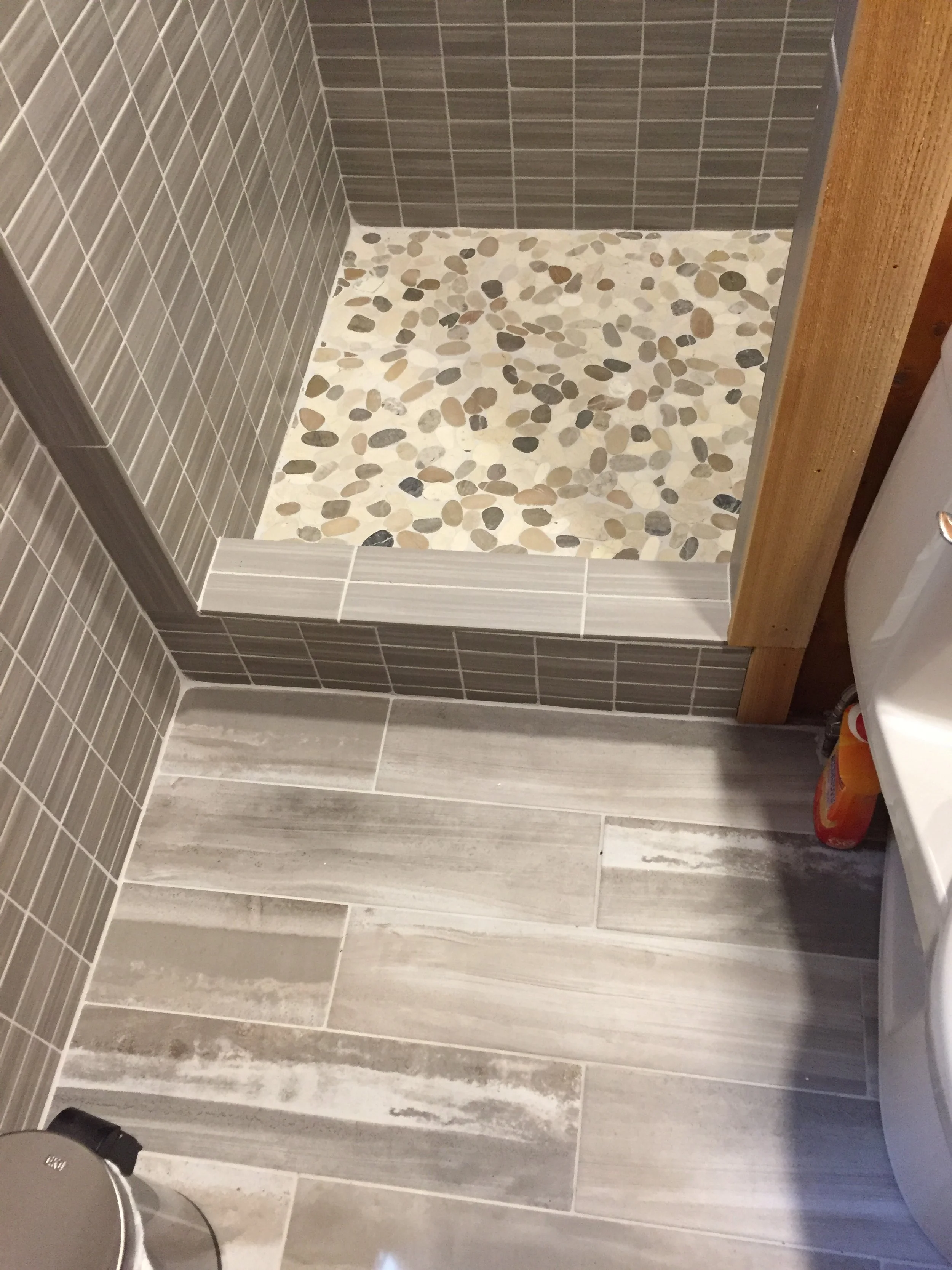 Bathroom with a pebble shower floor, gray tiled walls, and wood accent. Sliding bathroom door. Part of a washing machine and a detergent bottle visible.
