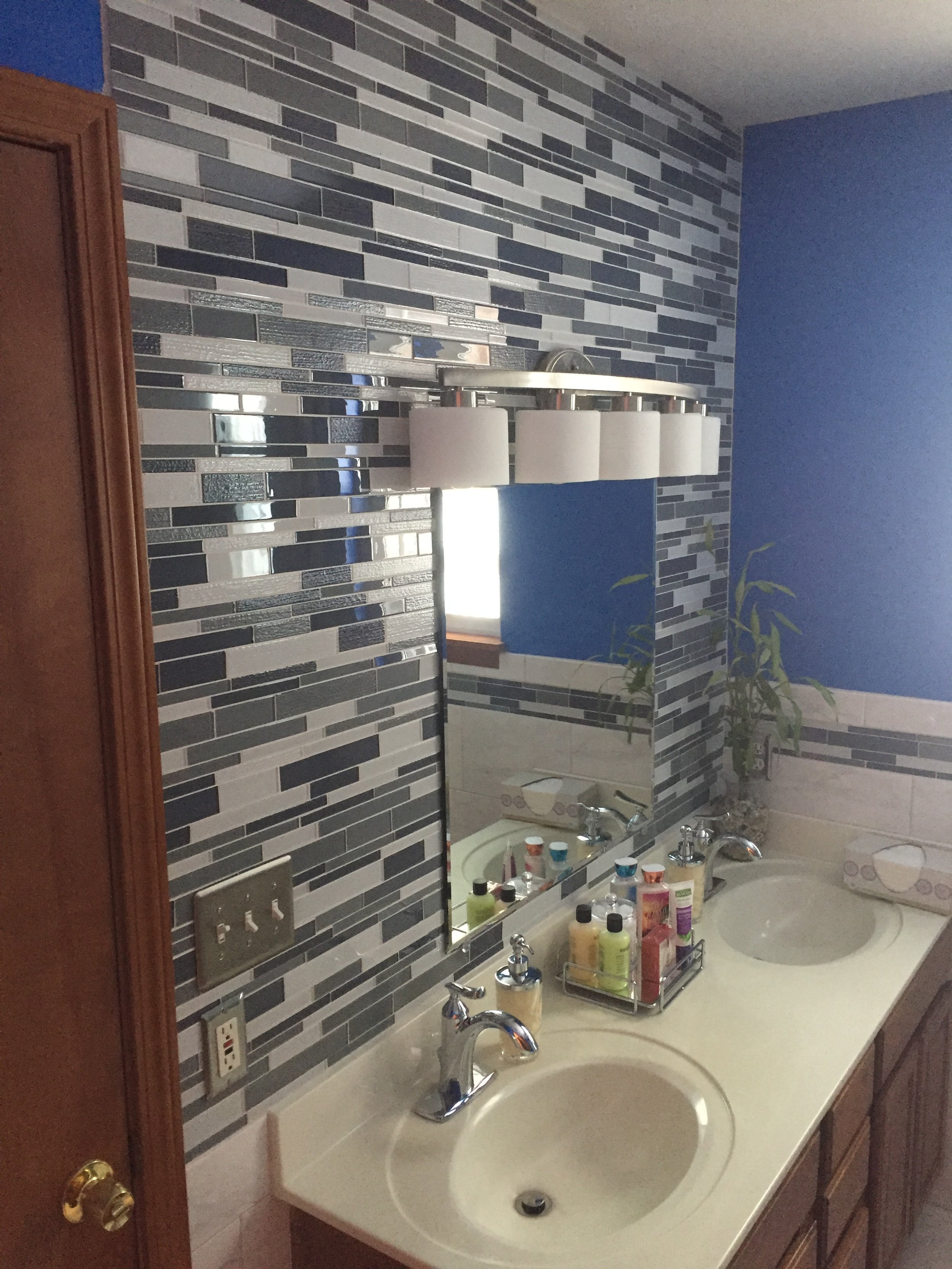 Bathroom vanity with two sinks, a large mirror, and a wall-mounted light fixture with five white cylindrical shades. The wall behind the vanity has a patterned tile design in shades of gray and blue. There is a small potted plant and various toiletri
