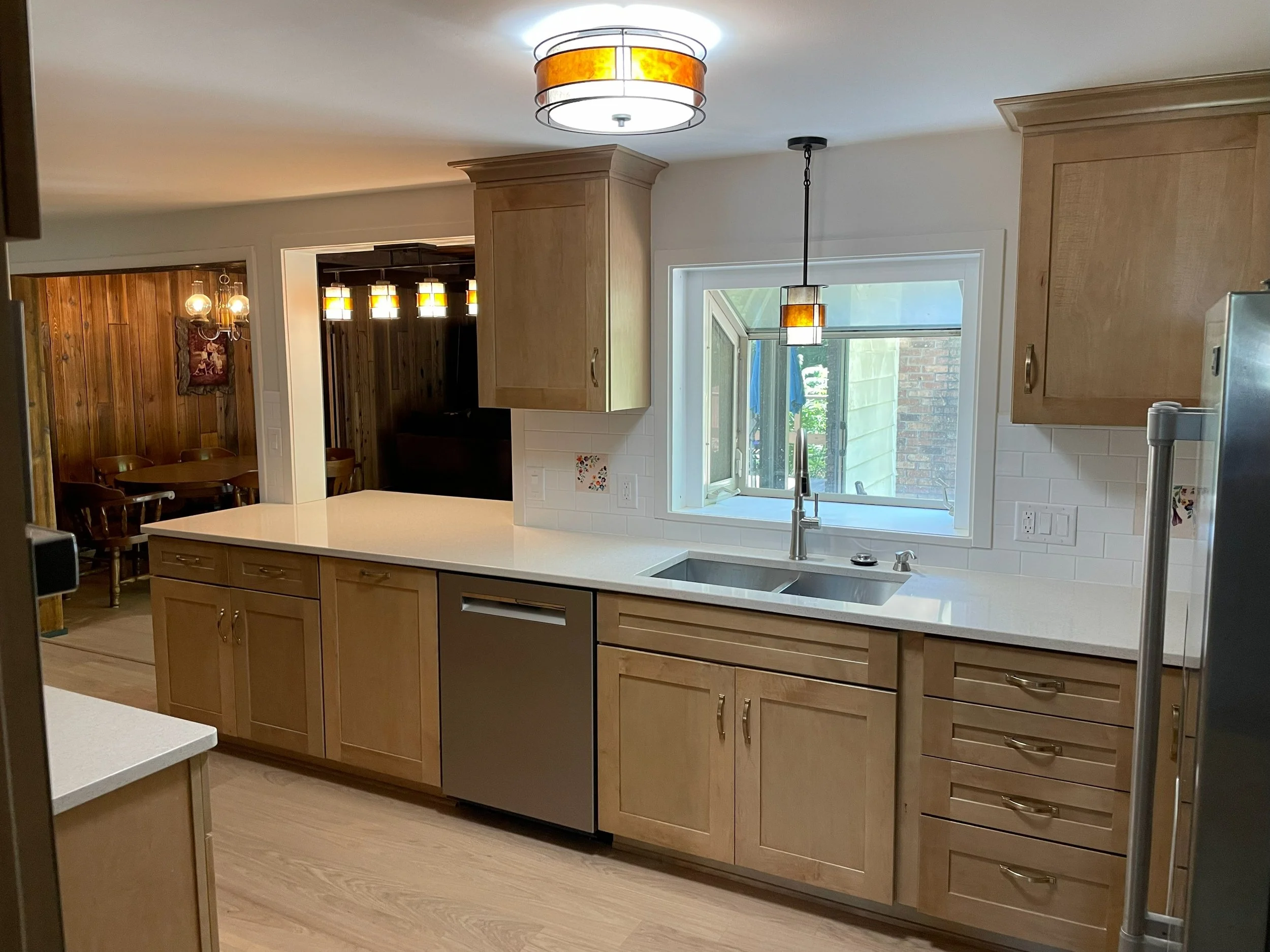 Kitchen with light wood cabinets, white countertop, double sink under a window, and stainless steel dishwasher. Adjacent dining area with wooden chairs and a wall-mounted art piece.