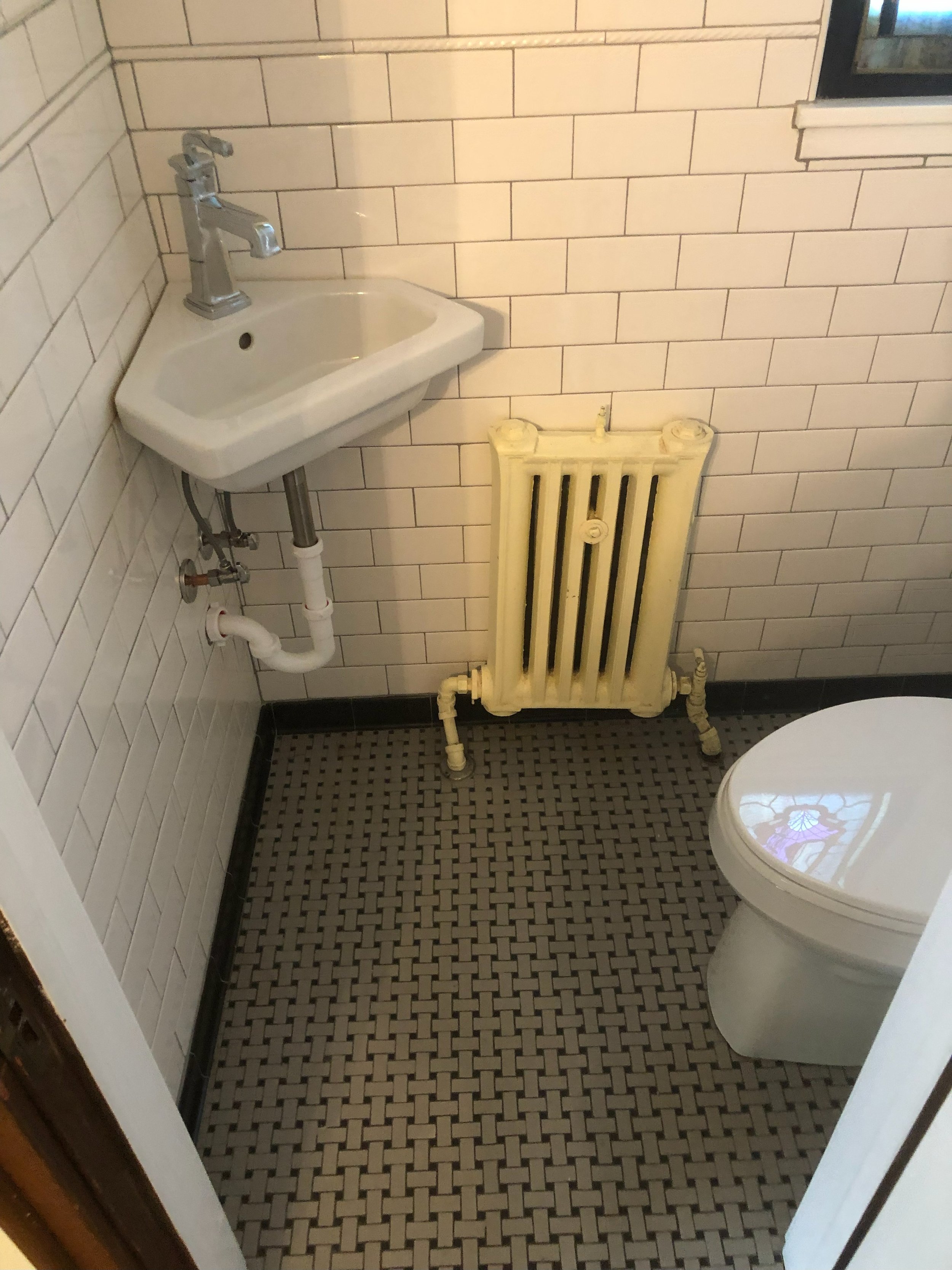 Small bathroom with white subway tile walls, a white wall-mounted sink with a chrome faucet, a vintage yellow radiator, and a toilet with a stained stained tank lid. The floor has a small black and white patterned tile design, and a small window is p