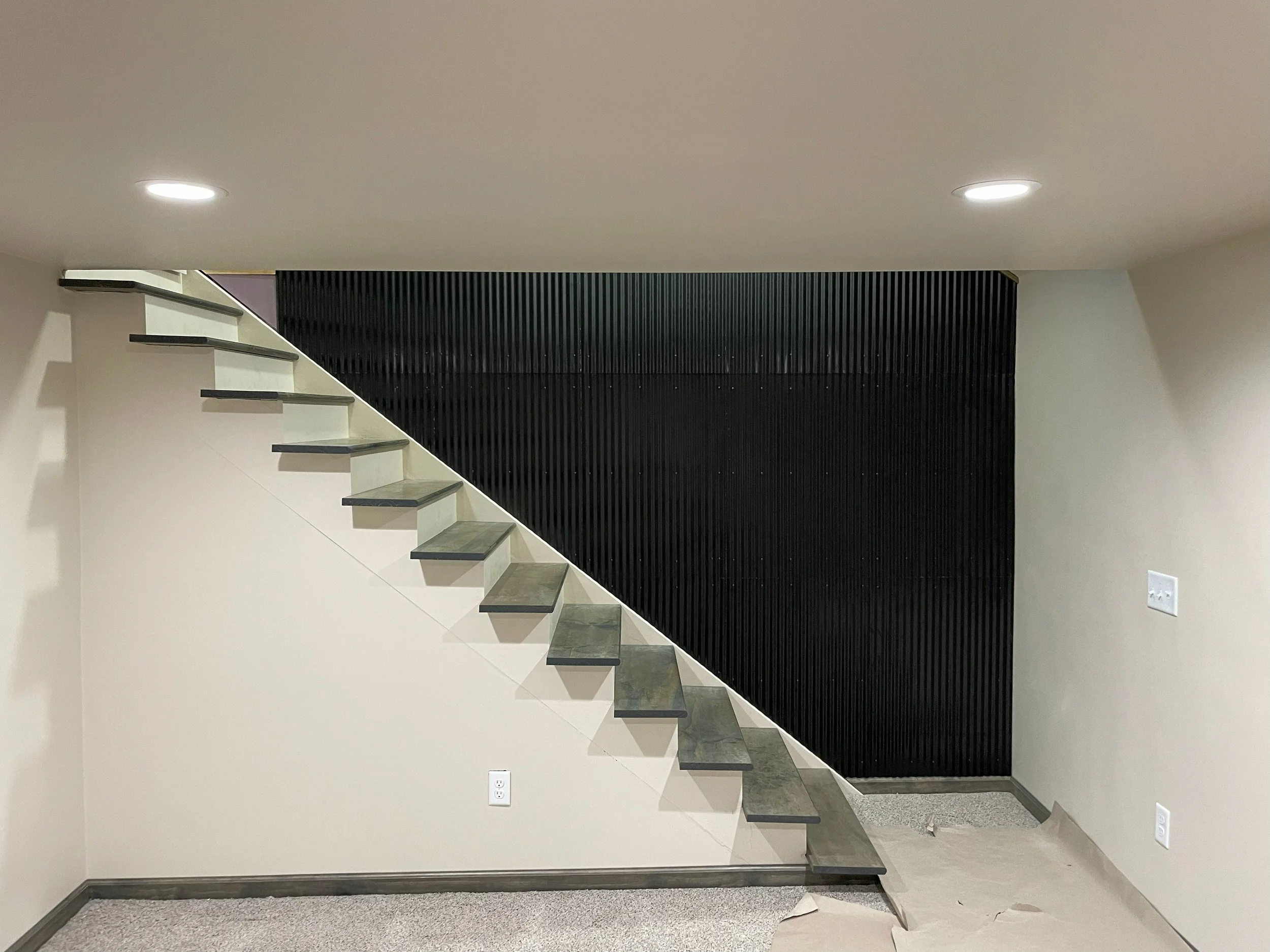 Interior view of a modern staircase with floating grey steps, black vertical slat wall behind it, and a white ceiling with two recessed lights.