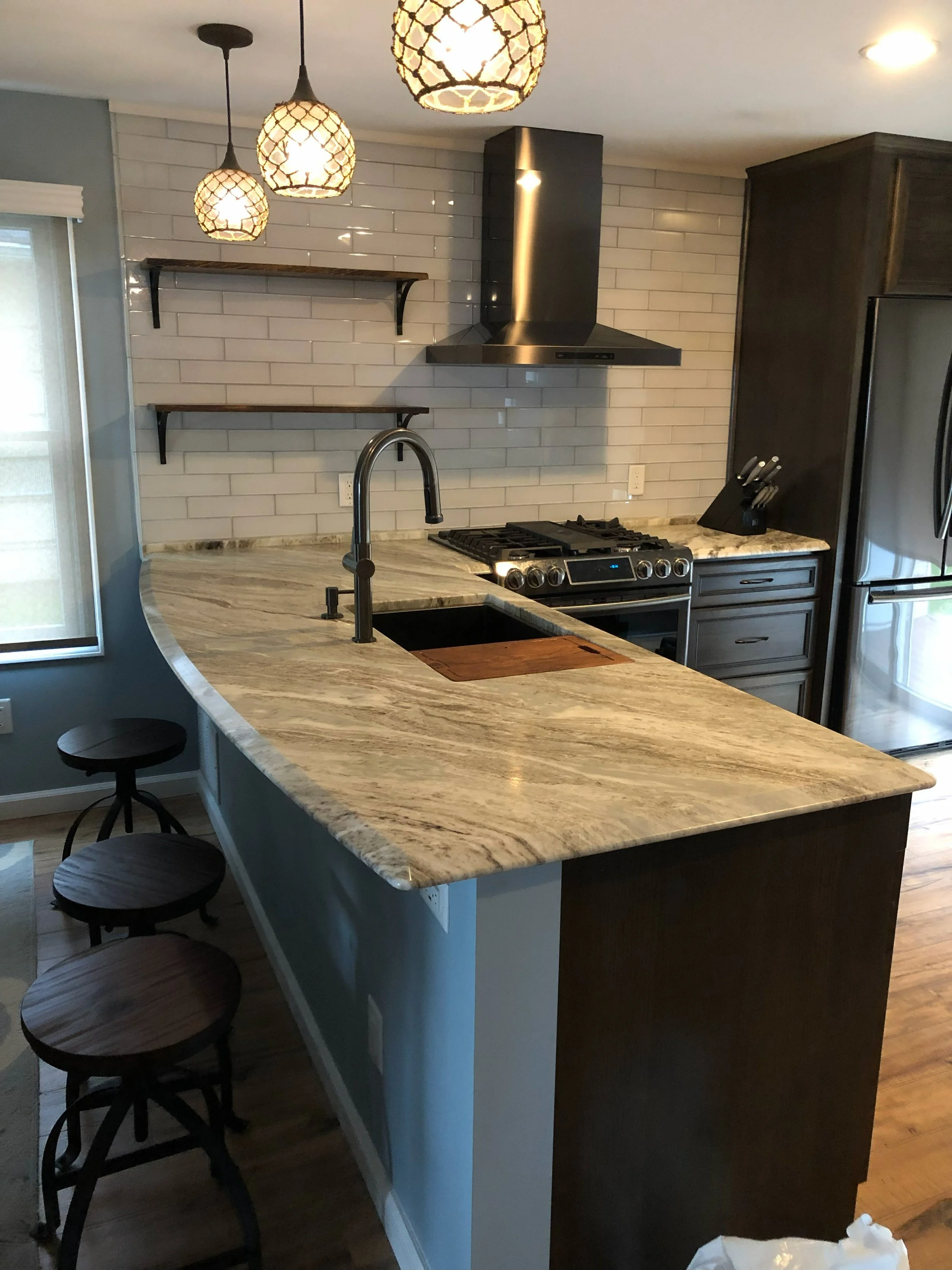 Kitchen with a curved granite countertop, pendant lights, white tile backsplash, stainless steel range hood and stove, dark cabinets, stainless refrigerator, window with blinds, and bar stools.