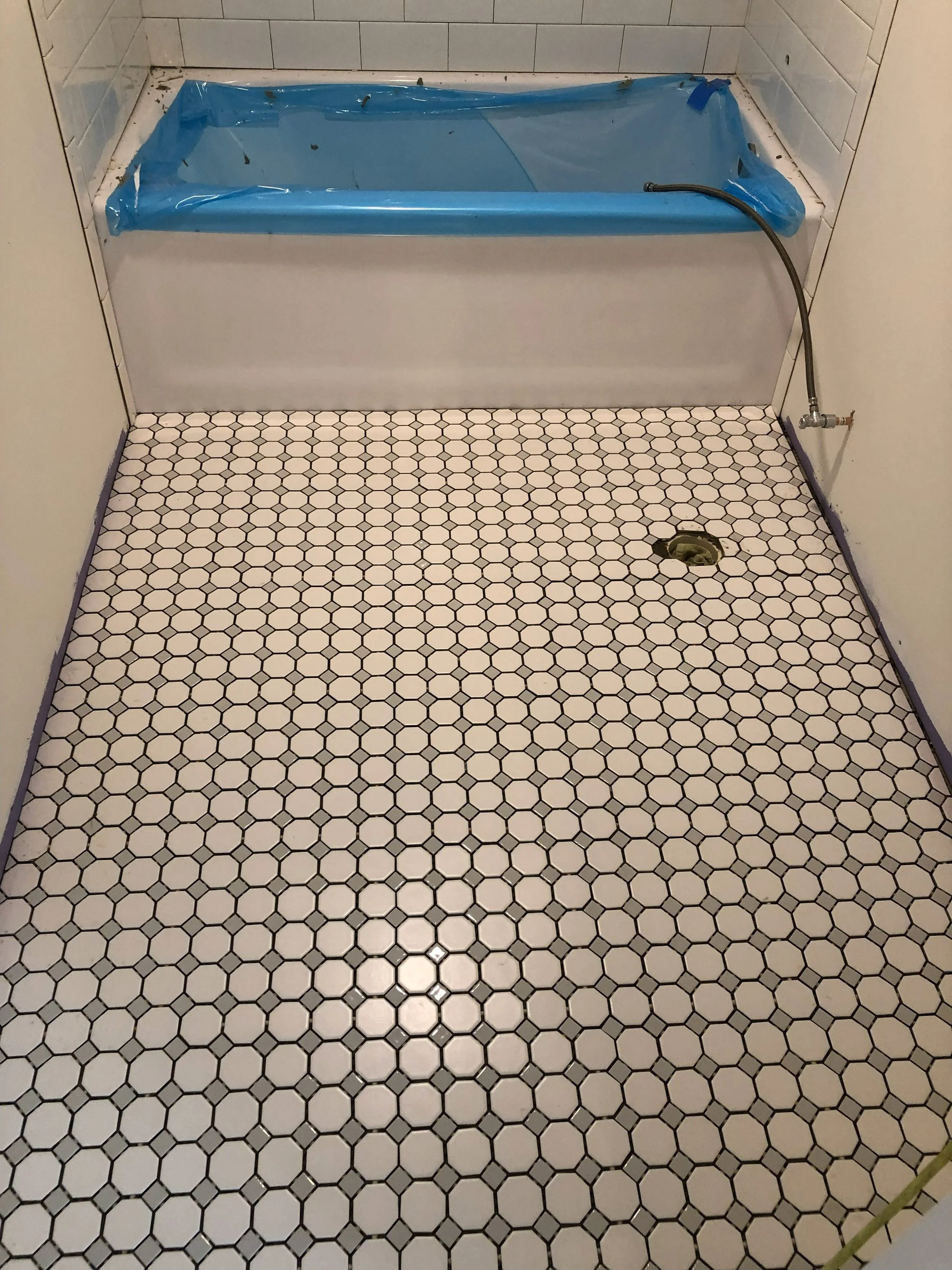 Bathroom with a hexagon-tile floor, a drain in the floor, and a bathtub covered with blue plastic and tape. There is also a hose connected to the bathtub faucet.