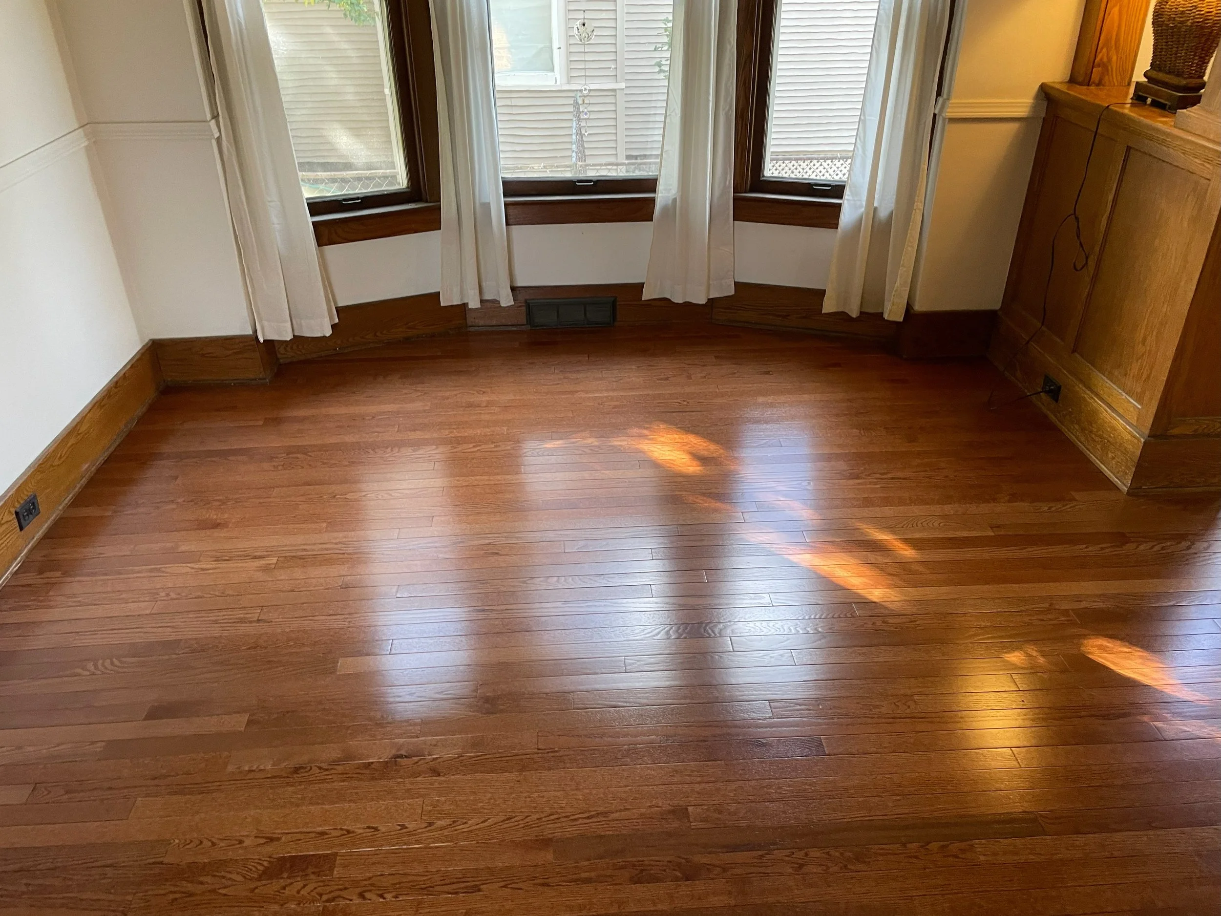 Empty room with wooden flooring and three windows with white curtains, sunlight reflecting on the floor.
