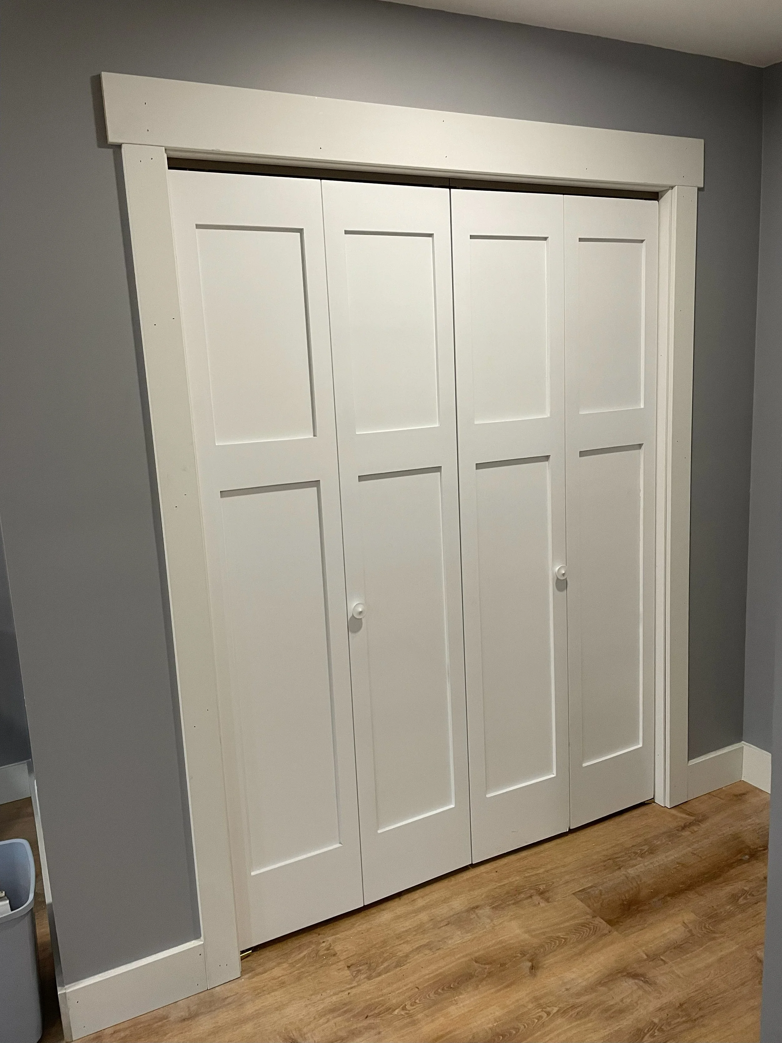 White door with 4 panels and a wooden frame around it, set in a gray wall with wooden flooring.