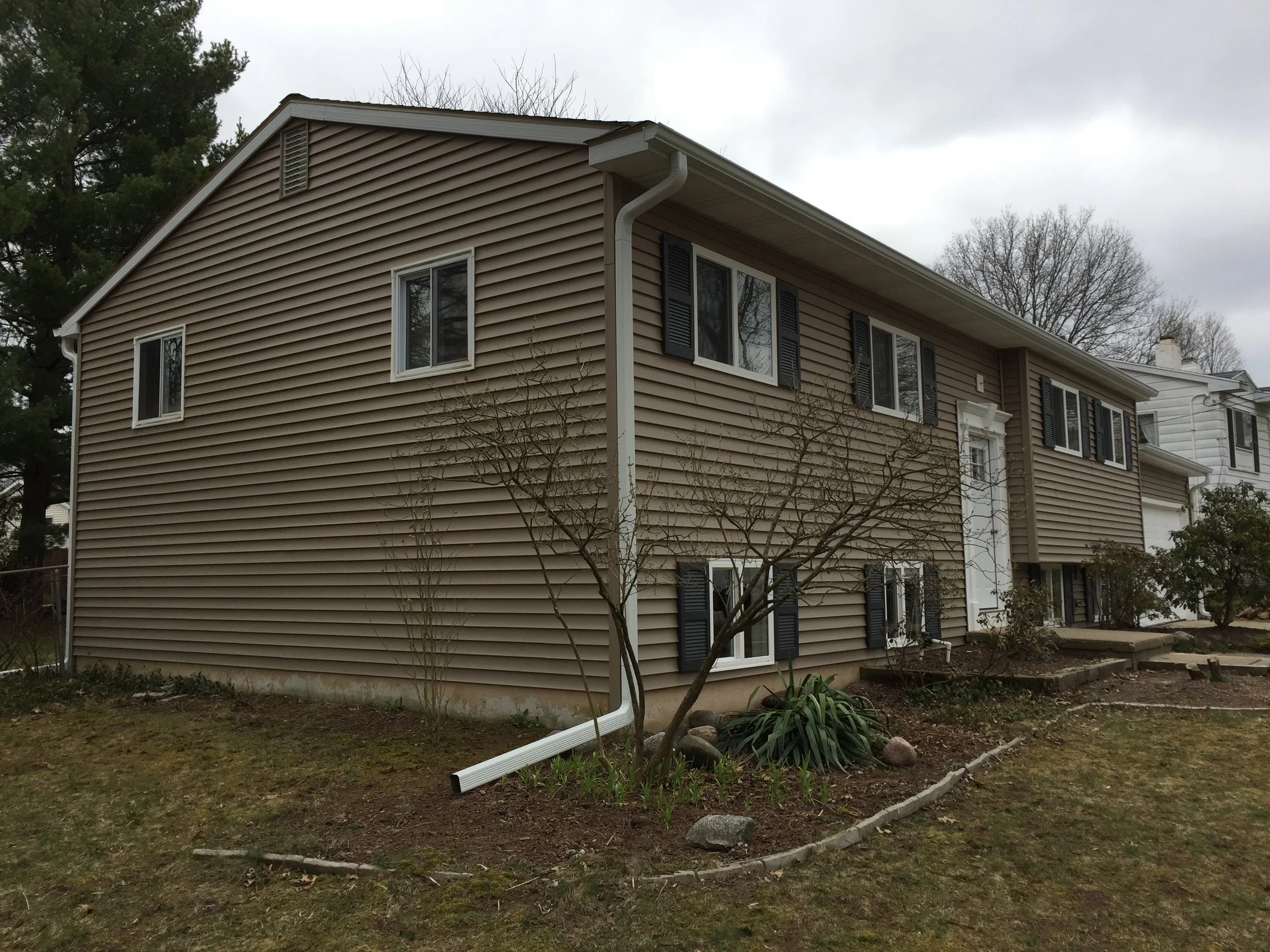 A two-story house with tan siding, white trim, and black shutters. The house has four windows on each floor and a white front door. There is a leafless tree and bushes in the front yard, with a garden bed and rocks around the base of the bushes. The 