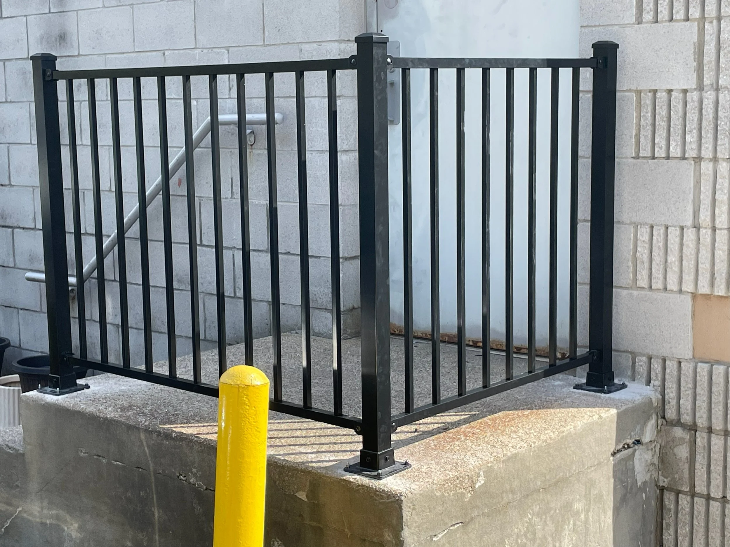 Black metal railing on a concrete platform with a yellow safety bollard in front, located outdoors near a building with gray brick walls and a metal door.