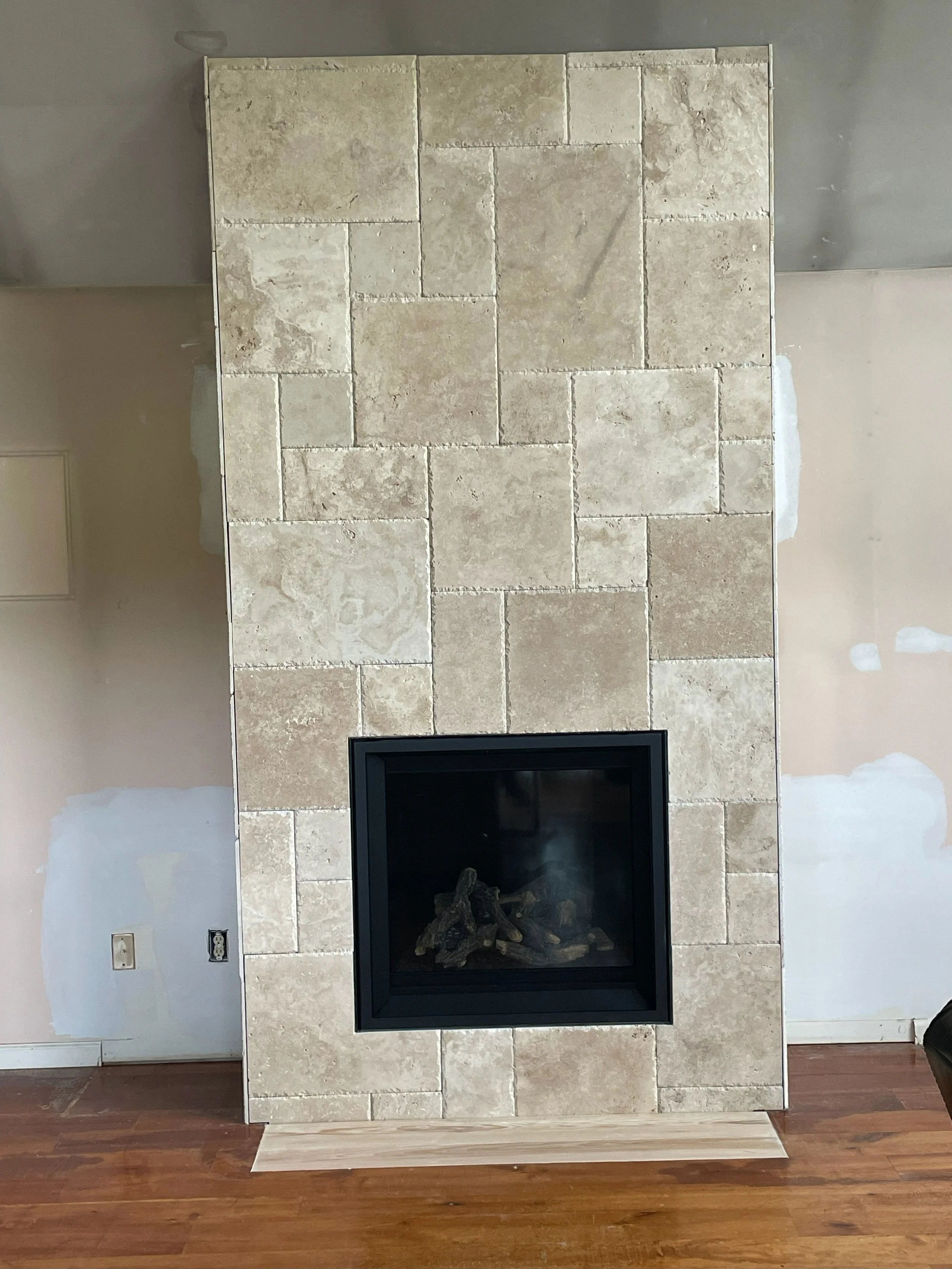 A beige stone fireplace with a black glass front, set against a wall with patches of unpainted drywall, on a hardwood floor.