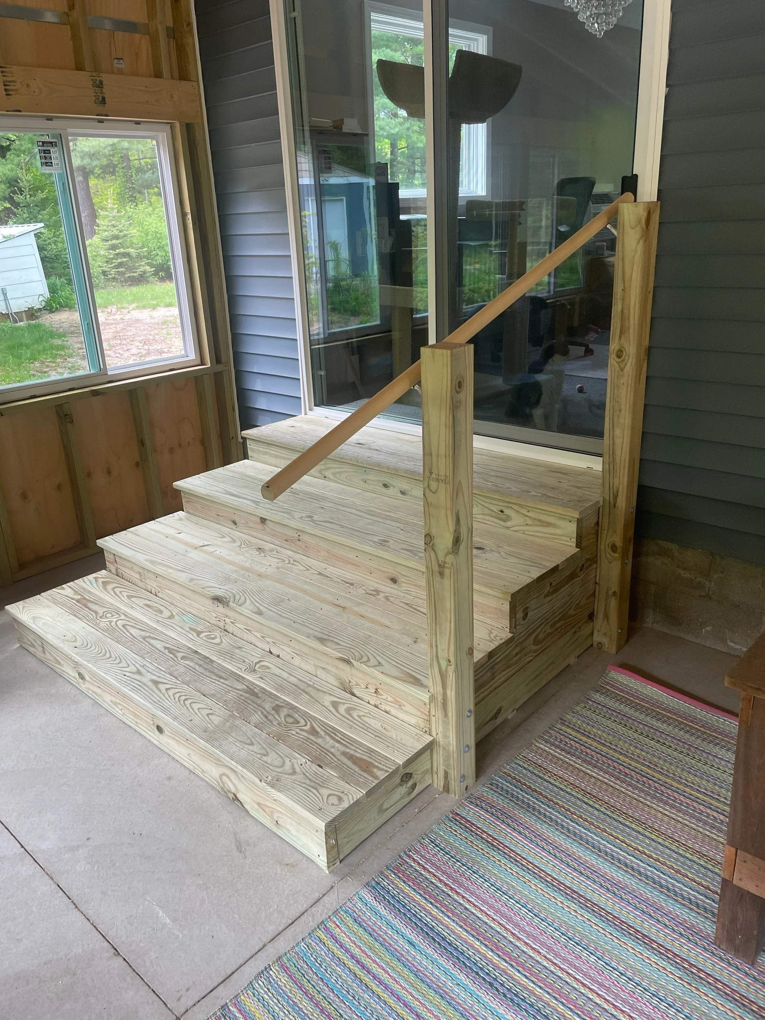 Unfinished wooden porch stairs with a handrail, leading up to a sliding glass door in a house under construction, with a window to the left and a colorful striped rug on the concrete floor.