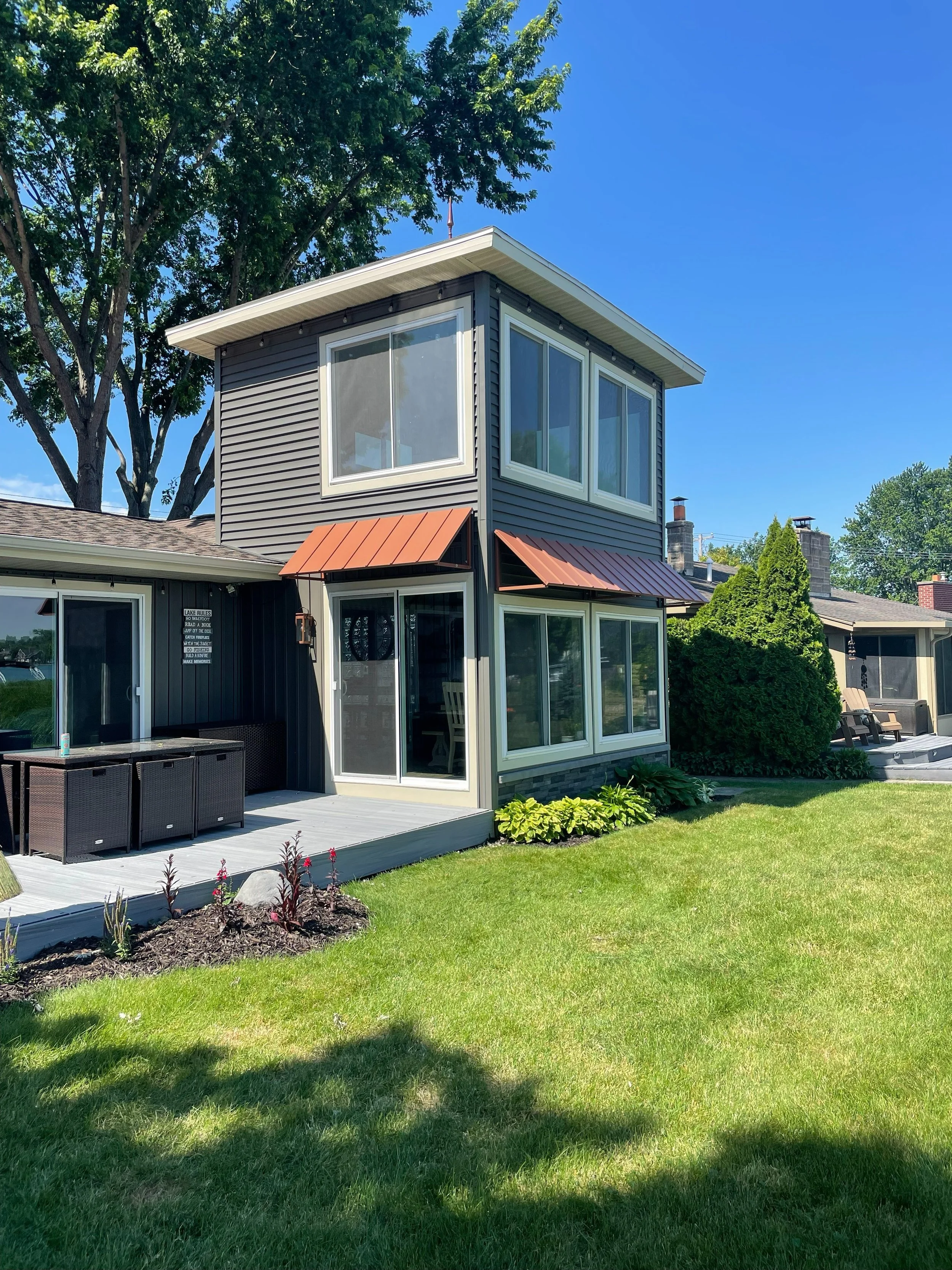 A modern two-story house with dark gray siding and large windows, surrounded by green grass, trees, and shrubs, with a small patio area.