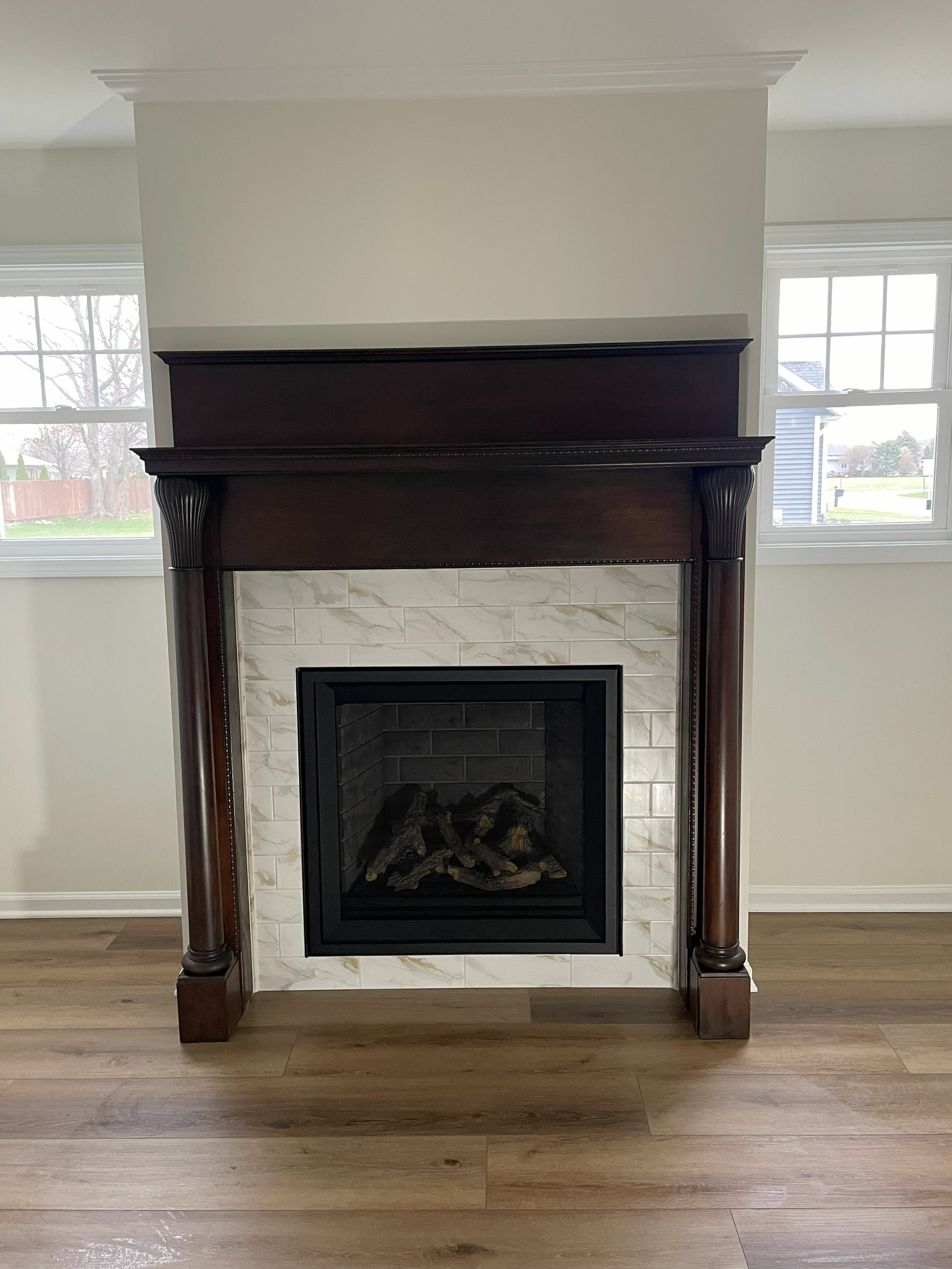 Fireplace with dark wooden mantel and marble tiles, situated in a bright room with two large windows and wood flooring.