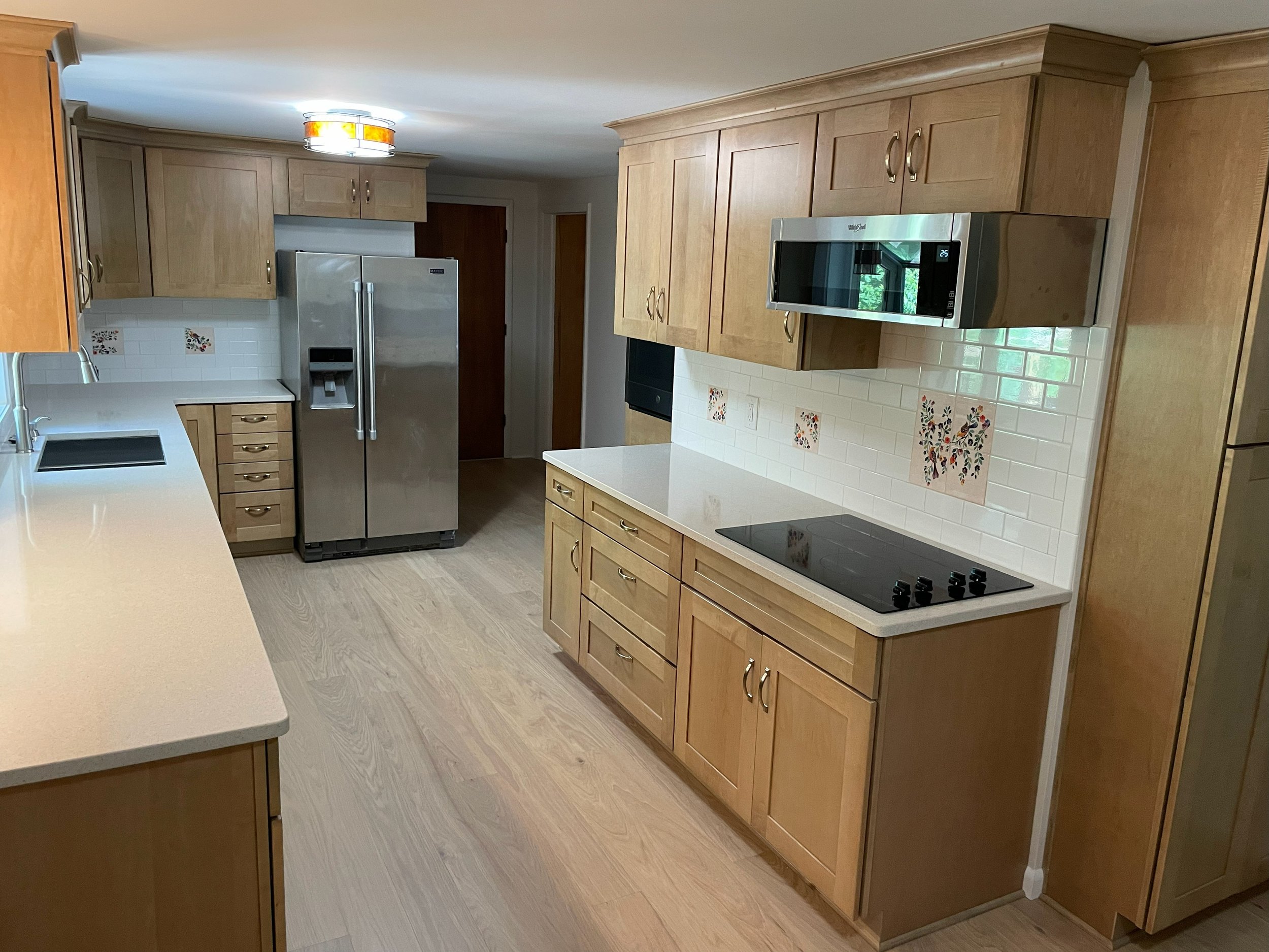 Kitchen with light wood cabinets, white countertops, a stainless steel refrigerator, a microwave, a ceramic cooktop, and a white tiled backsplash with decorative tiles.