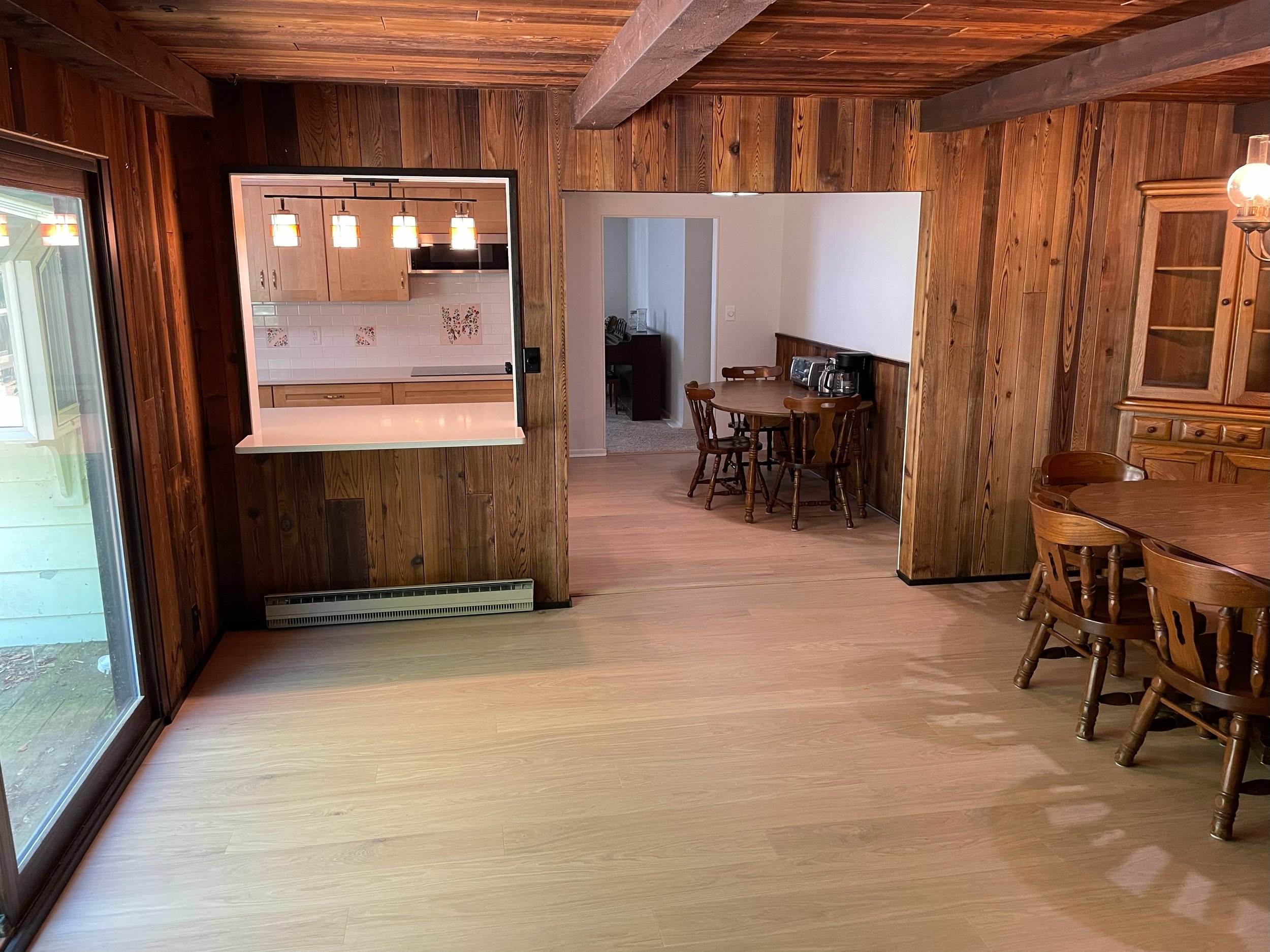 A dining area with wooden panel walls and furniture, a sliding glass door to the outside, and a view into the kitchen and adjacent room.