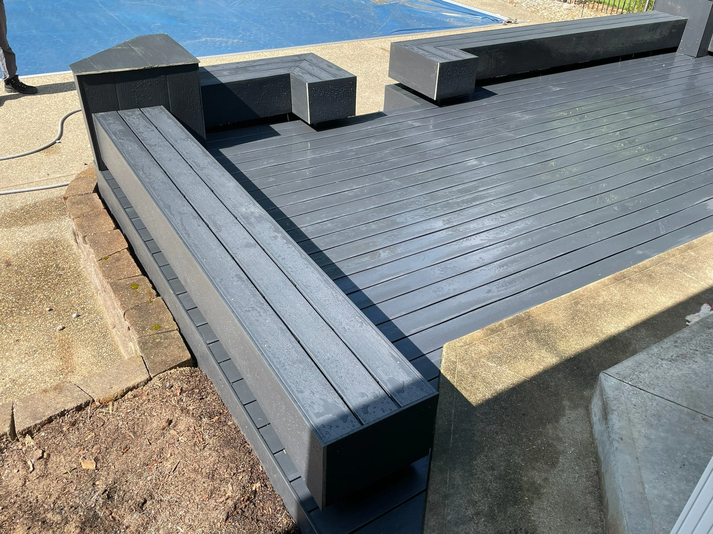 Black wooden poolside seating area with benches and a platform, adjacent to a swimming pool, with a concrete and brick border and some dirt and grass visible nearby.