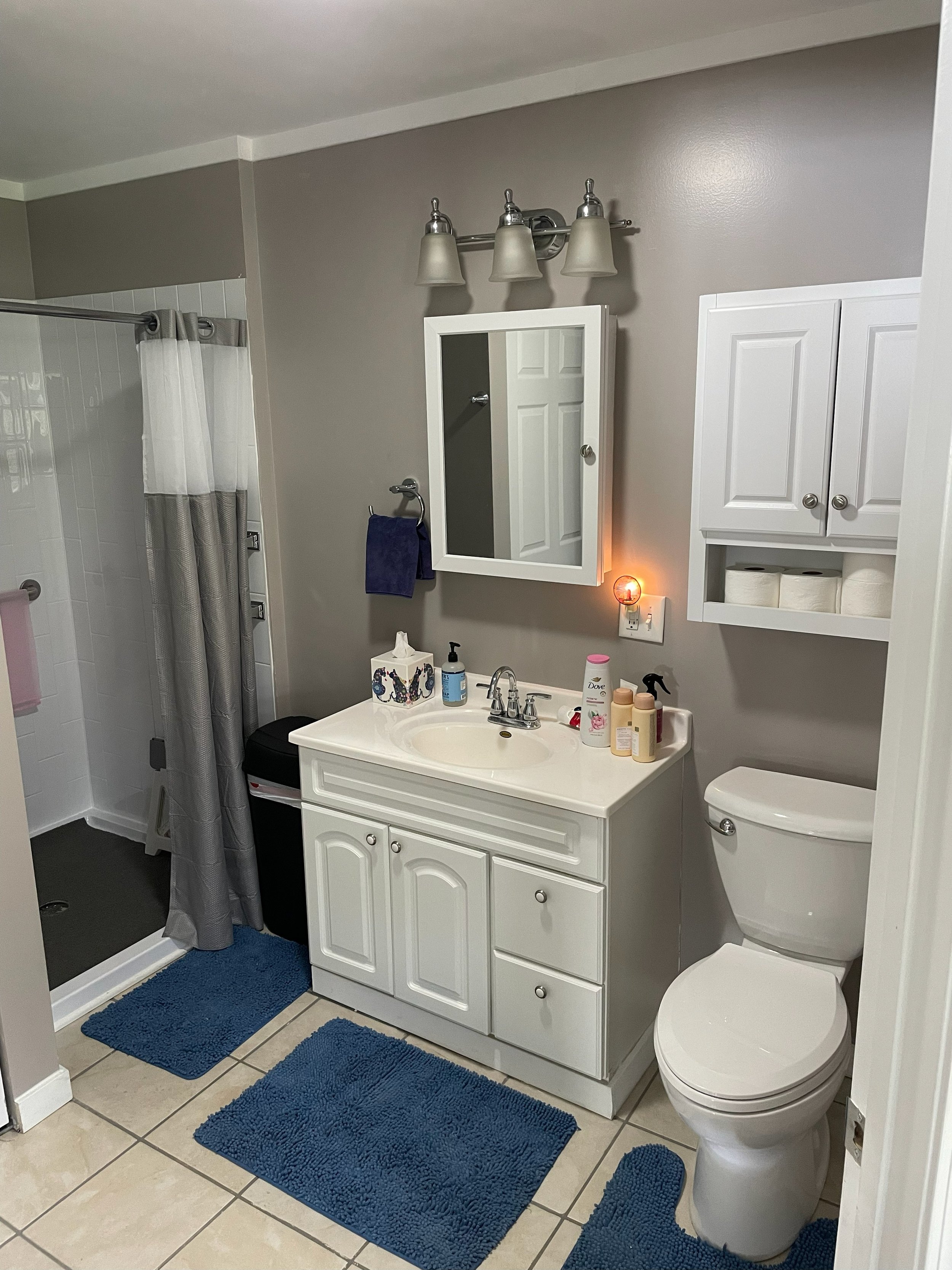 A small bathroom with a shower, white vanity with a mirror, cabinets, toilet, blue rugs, and wall-mounted light fixture.