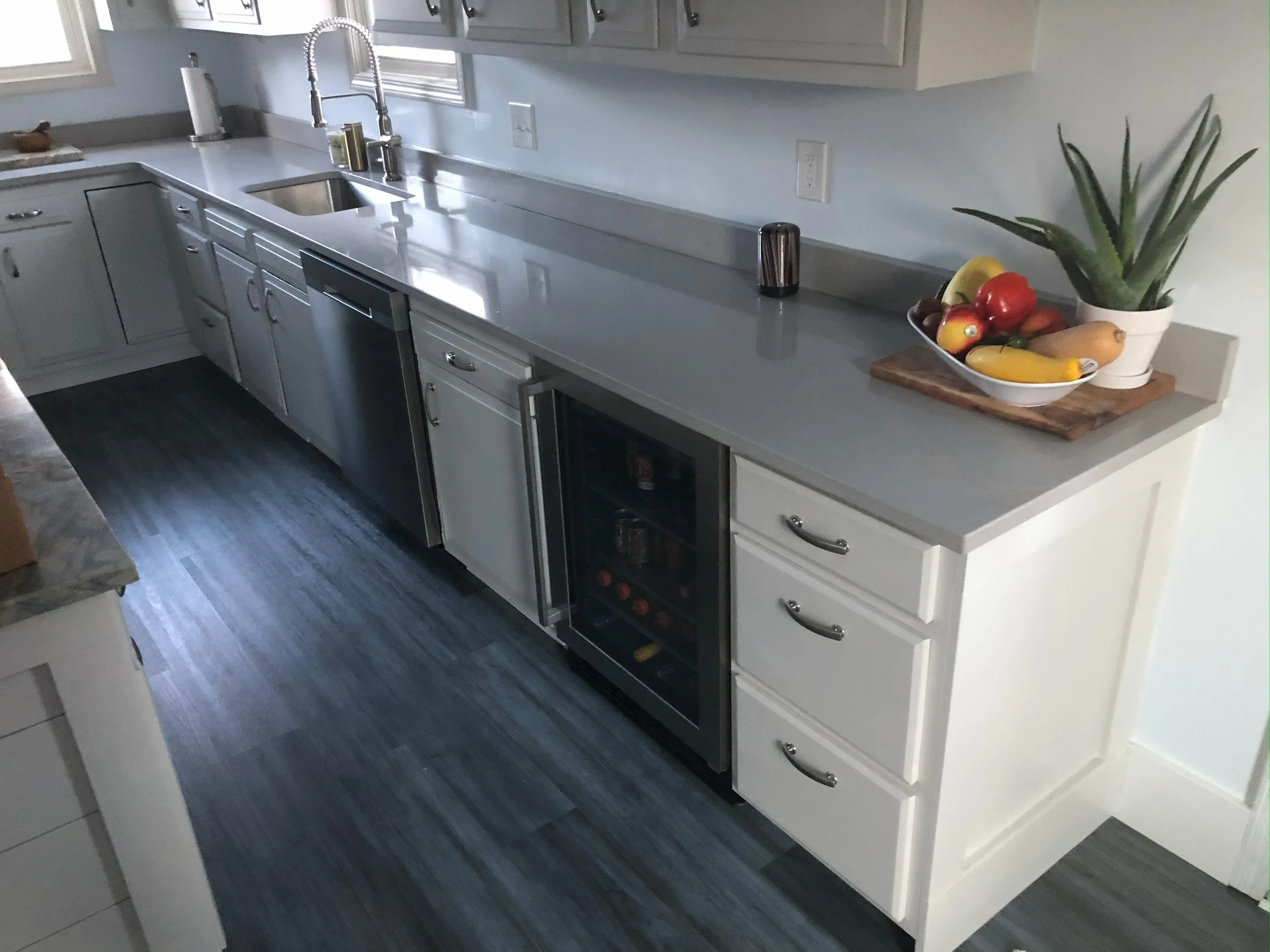 Modern kitchen with gray countertop, white cabinets, stainless steel sink, small wine fridge, fruit bowl, and aloe vera plant.