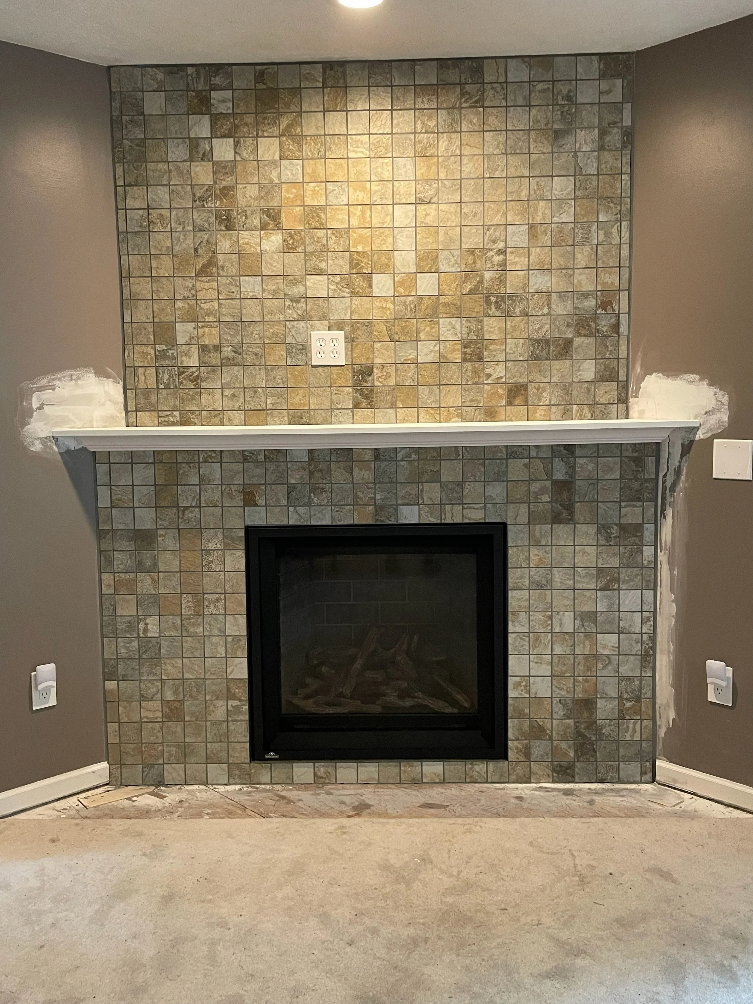 Newly renovated fireplace with tile surround and brown walls, with unfinished flooring and electrical outlets.