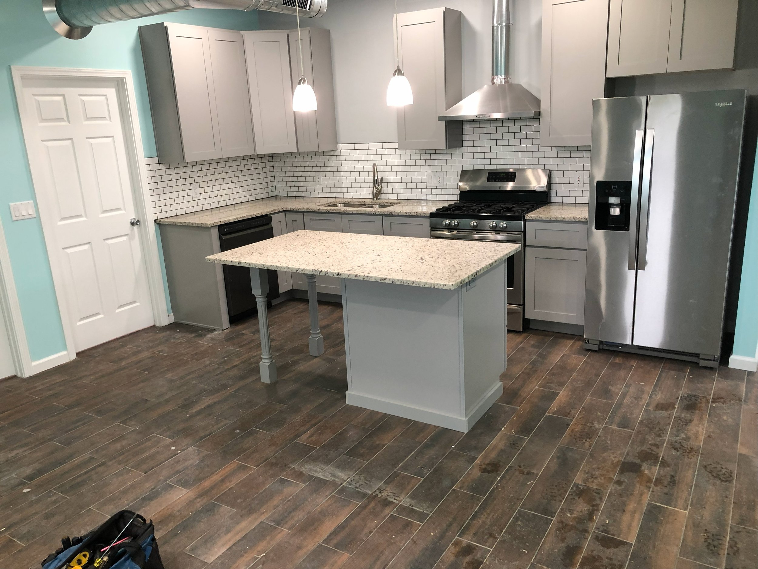 Modern kitchen with gray cabinets, white subway tile backsplash, granite countertops, and stainless steel appliances, including a refrigerator, oven, and range hood. There is a small island with a granite top, pendant lighting, and wood-look tile flo