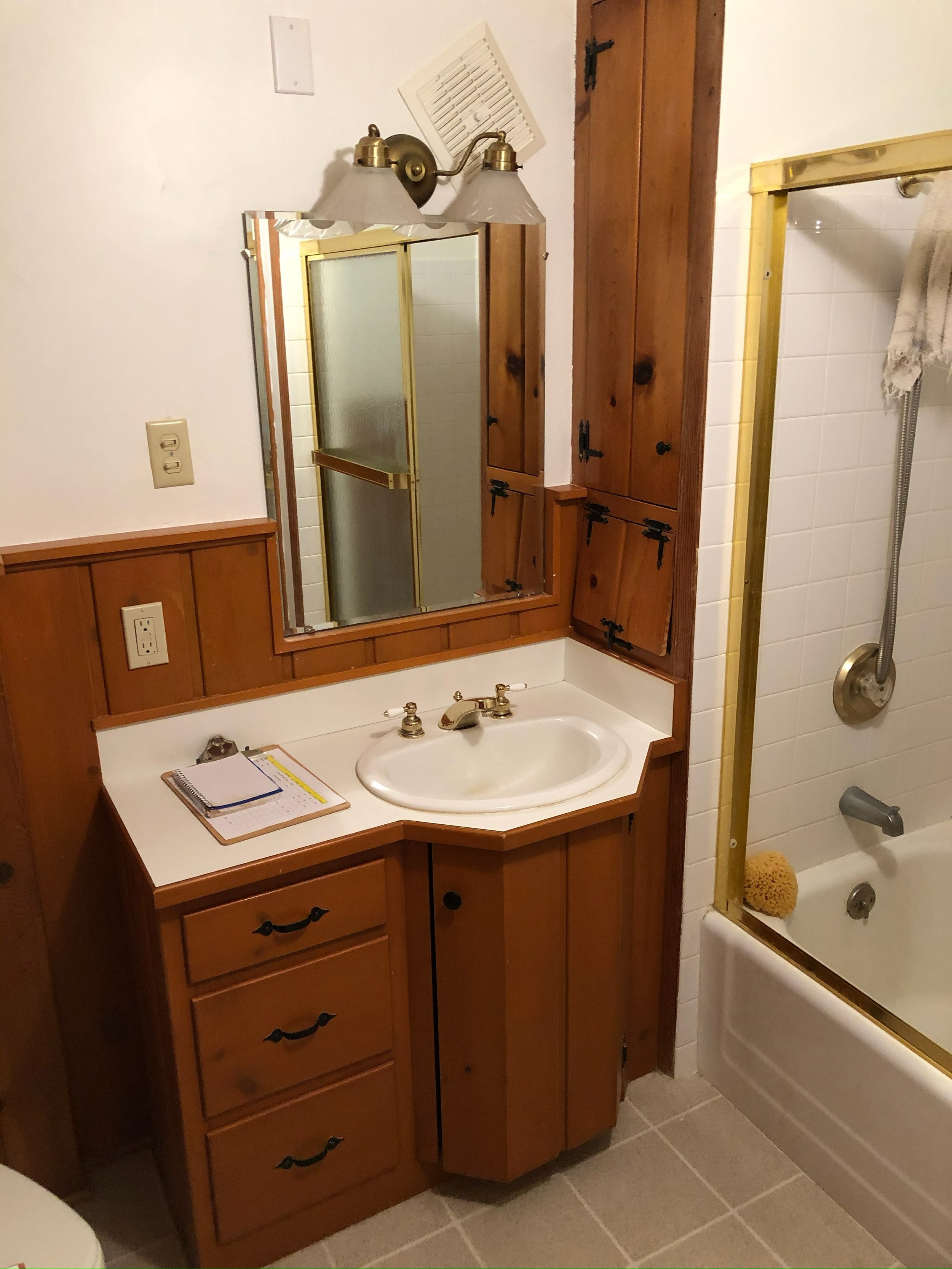 Bathroom with a wooden vanity, mirror, wall-mounted light fixture, small cabinet, and a shower with a glass door.
