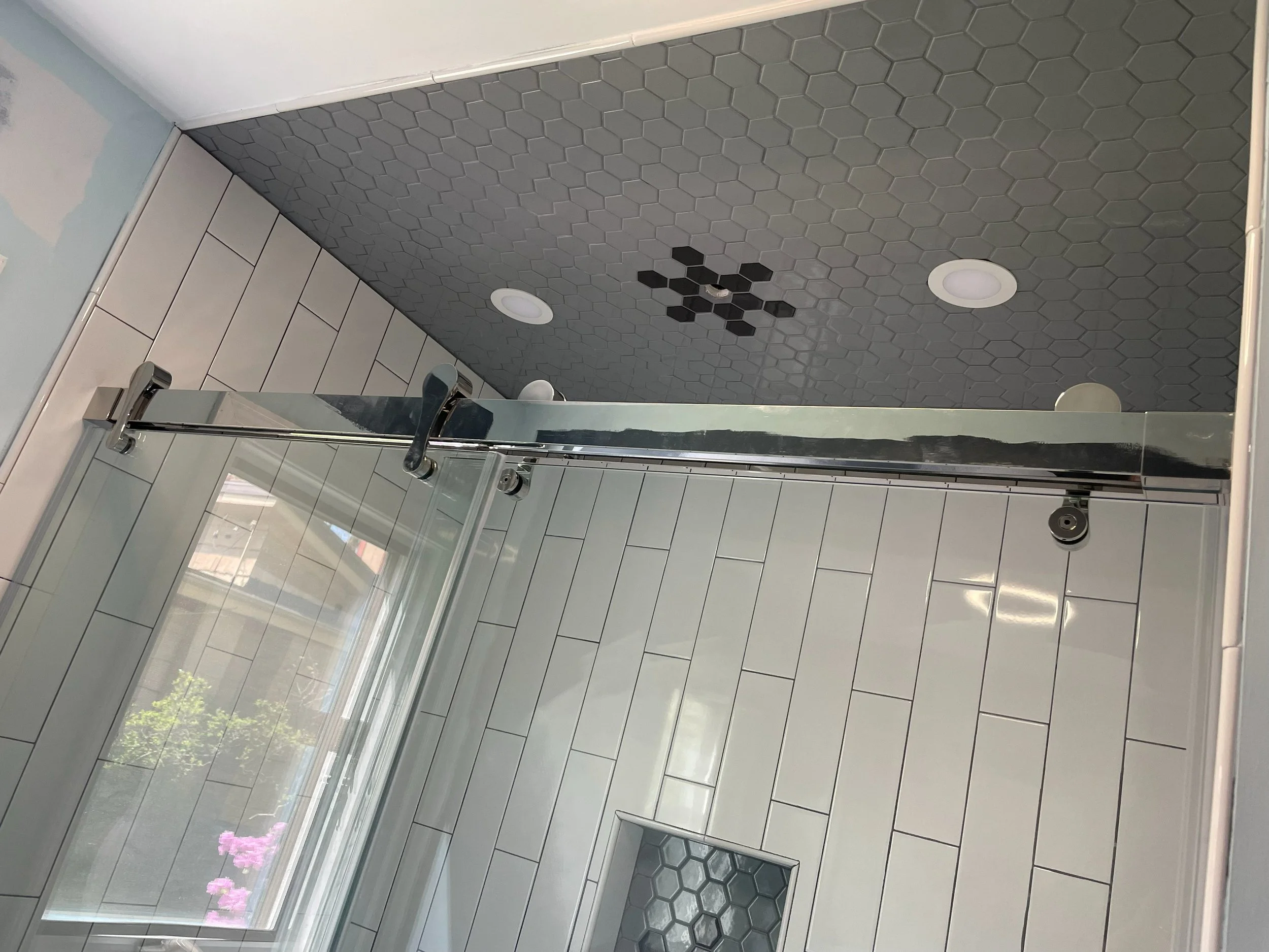 Bathroom shower area with a glass sliding door, white rectangular wall tiles, and a ceiling with dark hexagonal tiles and several circular recessed lights.