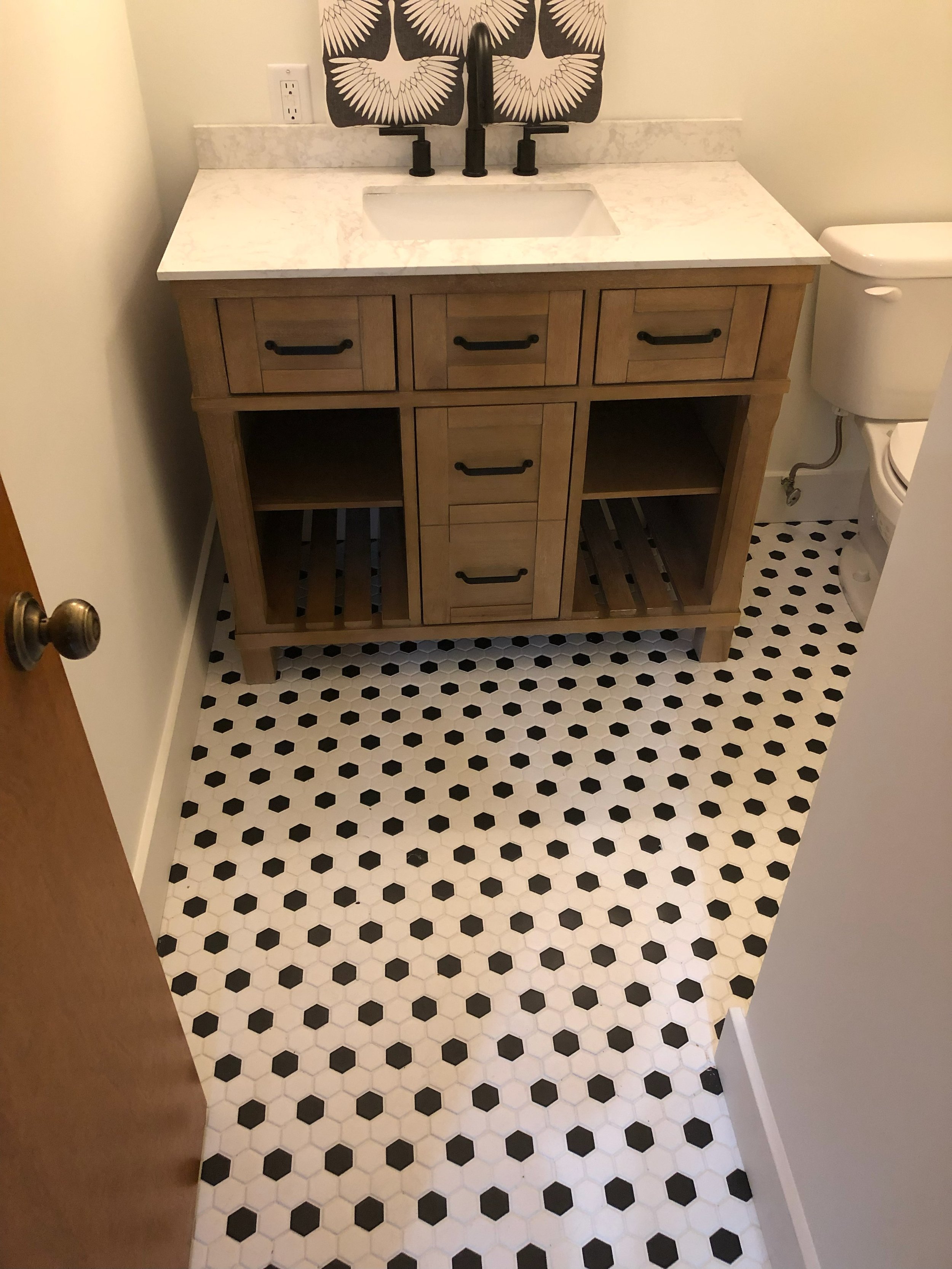 Bathroom with a wooden vanity with a white marble countertop, black hardware, and decorative backsplash, next to a white toilet, and black-and-white hexagon tile flooring.