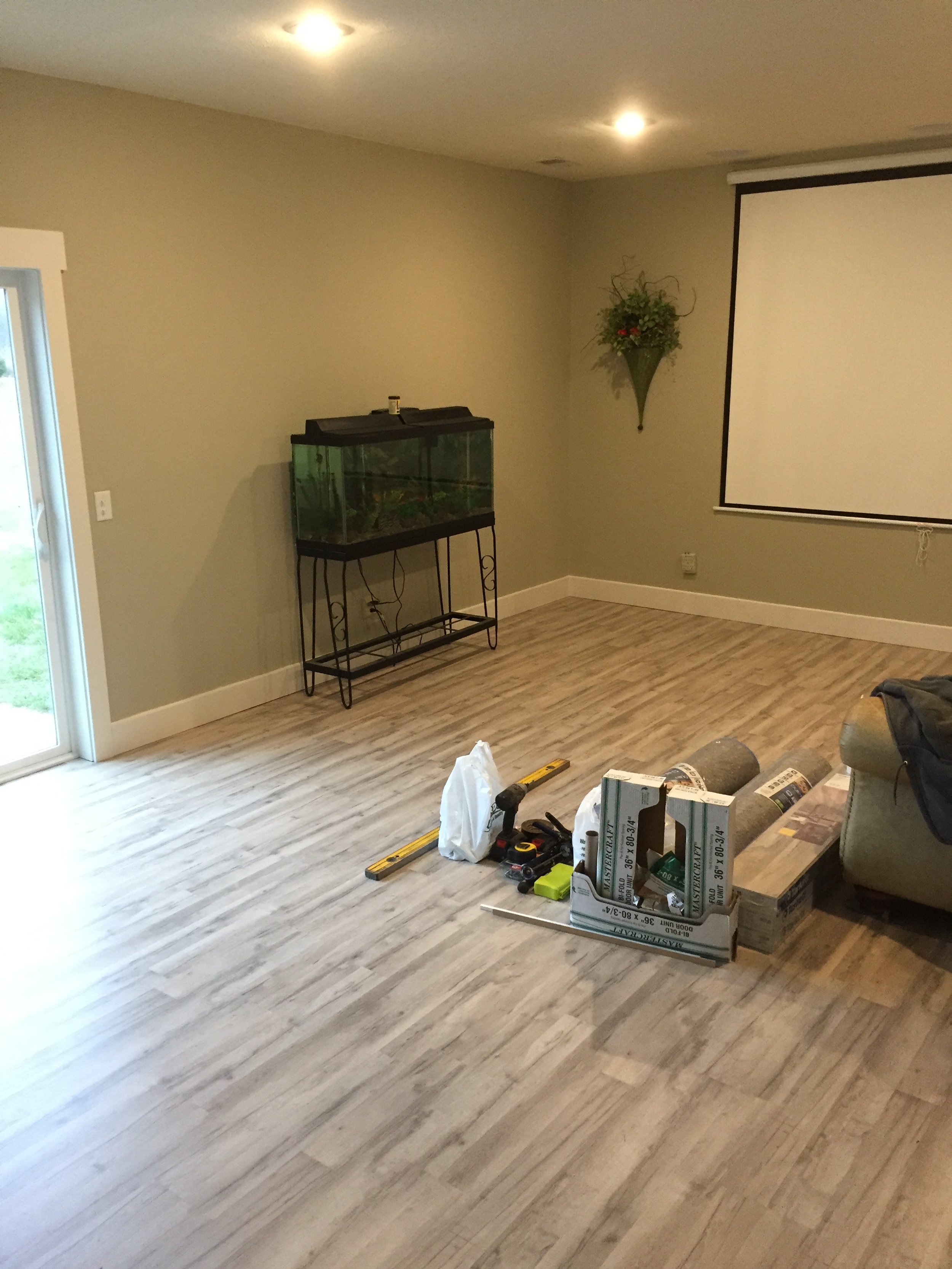 Empty living room with light wood flooring, a sliding glass door on the left, and a projector screen on the right. There are construction tools and materials on the floor, and a fish tank on a stand against the back wall.