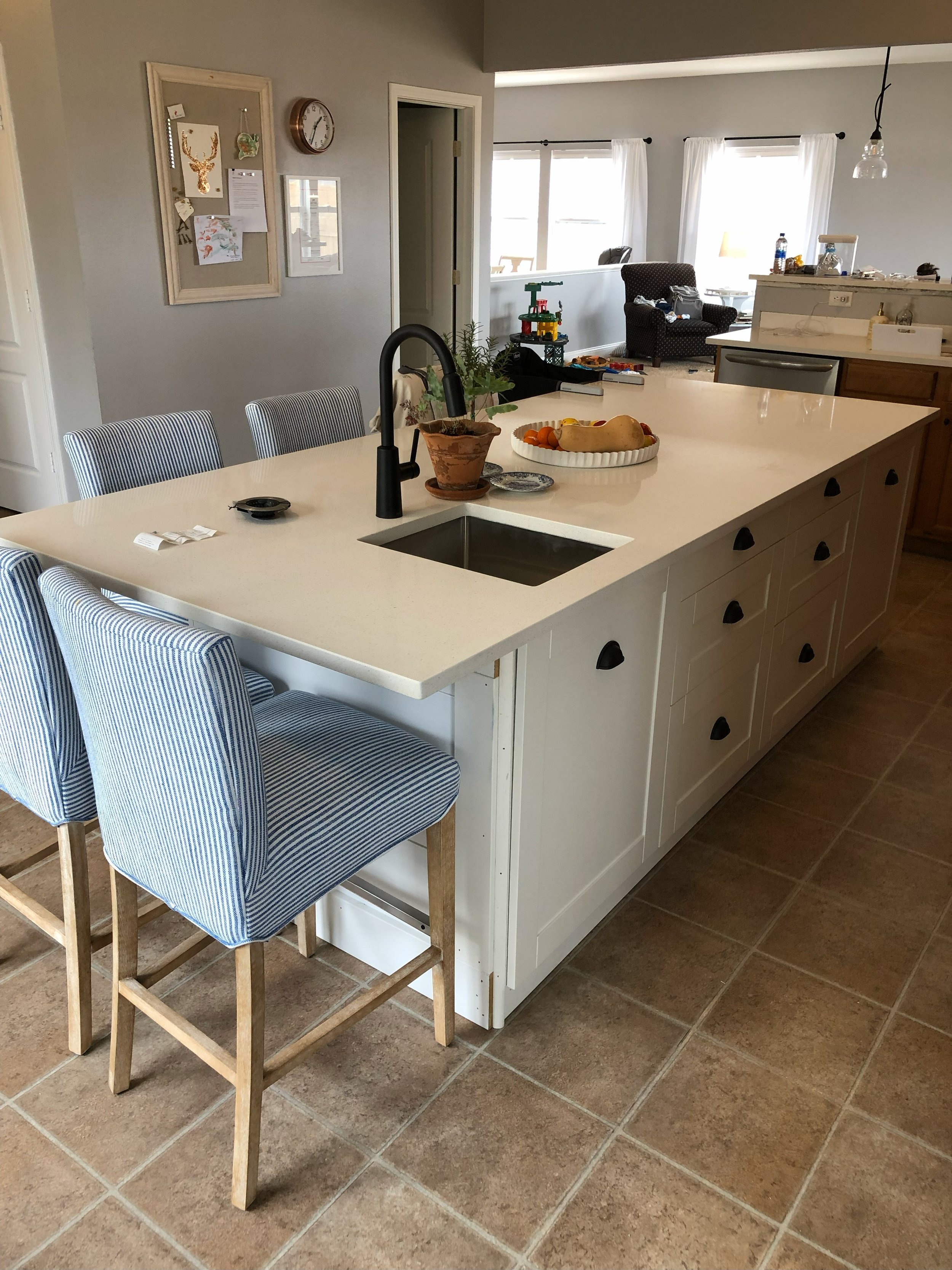 Kitchen island with a plant, fruit bowl, and a kitchen sink, with barstools on the side. Background shows a living room with armchair, toys, and windows with curtains.