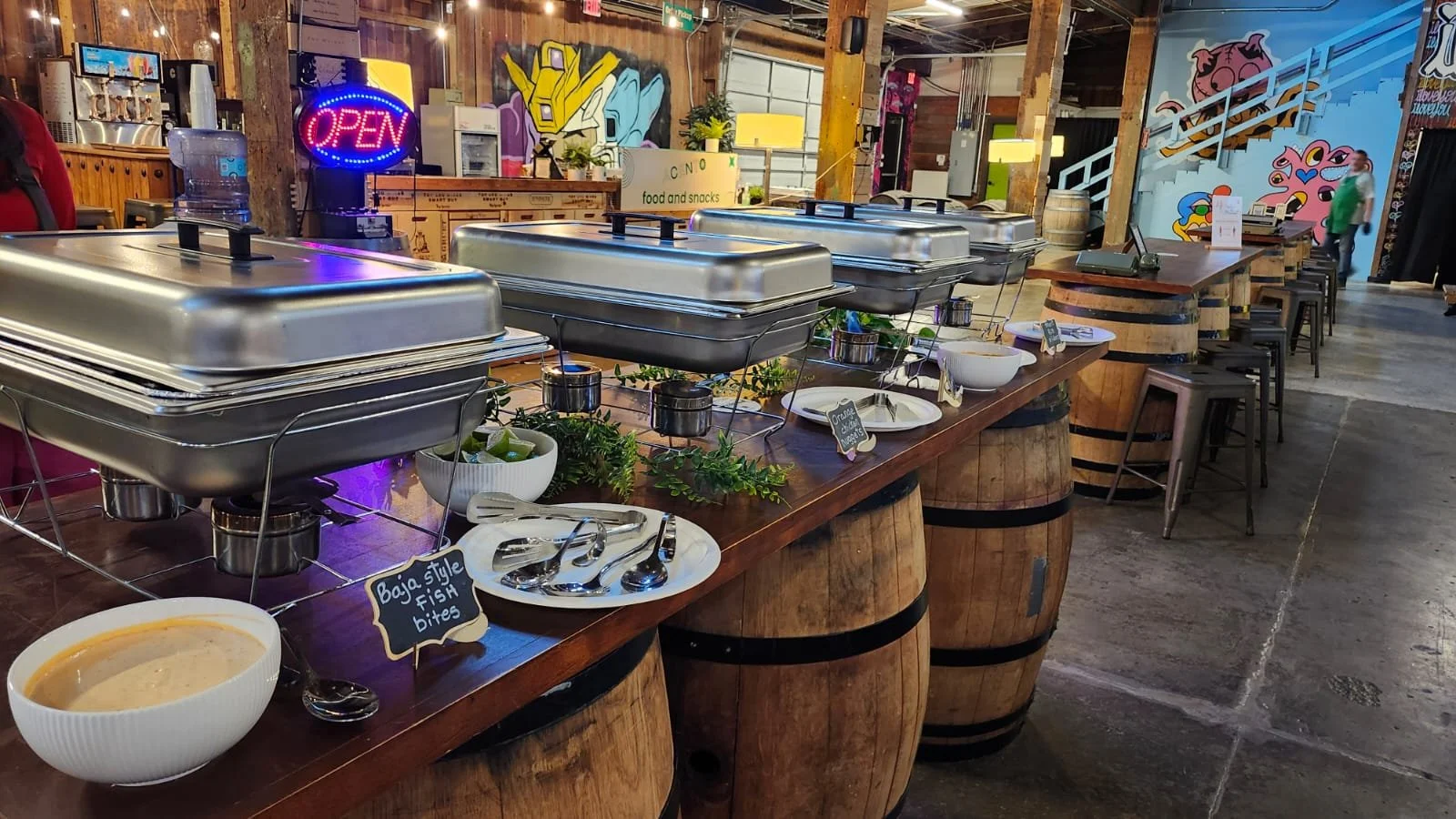 Rustic food presentation on barrel tables with chafing dishes against a backdrop of neon signs and graffiti art by Acento ATX