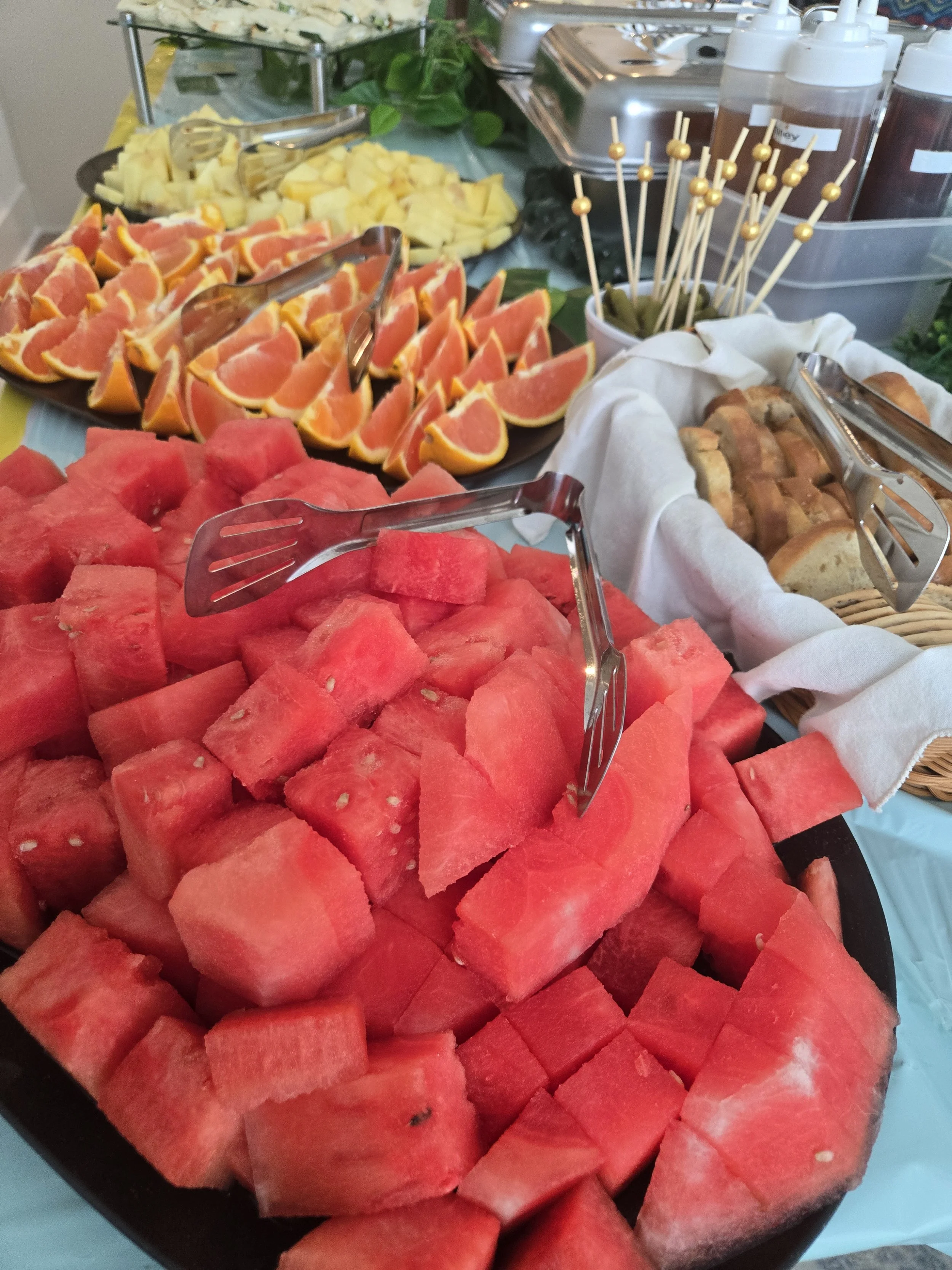 Close-up of a large black tray filled with cut watermelon cubes, with other fruits and snacks visible in the background like orange slices, pineapple, and bread rolls at a buffet table.