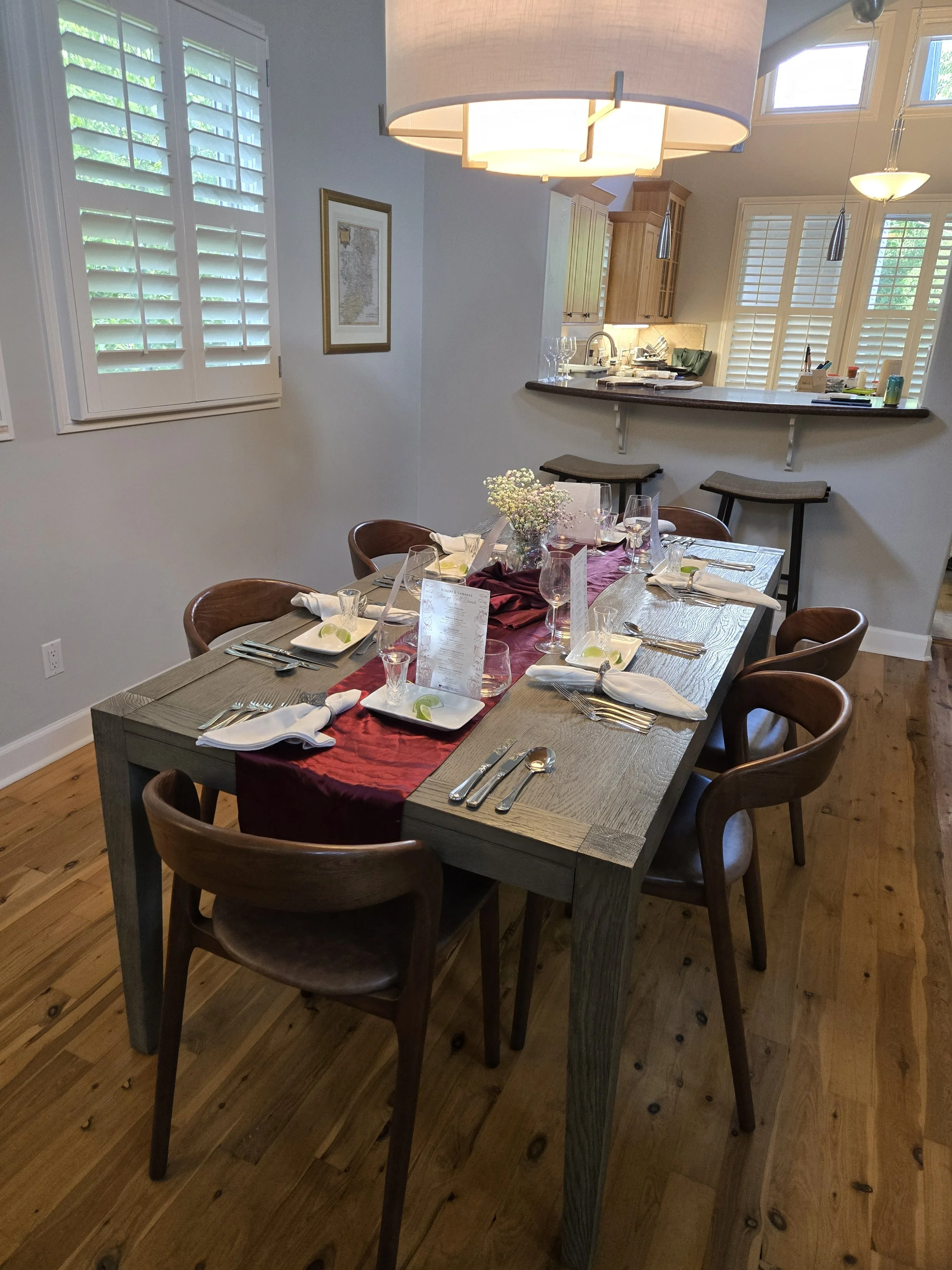 A dining room with a wooden table set with plates, glasses, silverware, and napkins, and a centerpiece of small white flowers. The room has large windows with white plantation shutters, hardwood flooring, and a hanging light fixture above the table. 