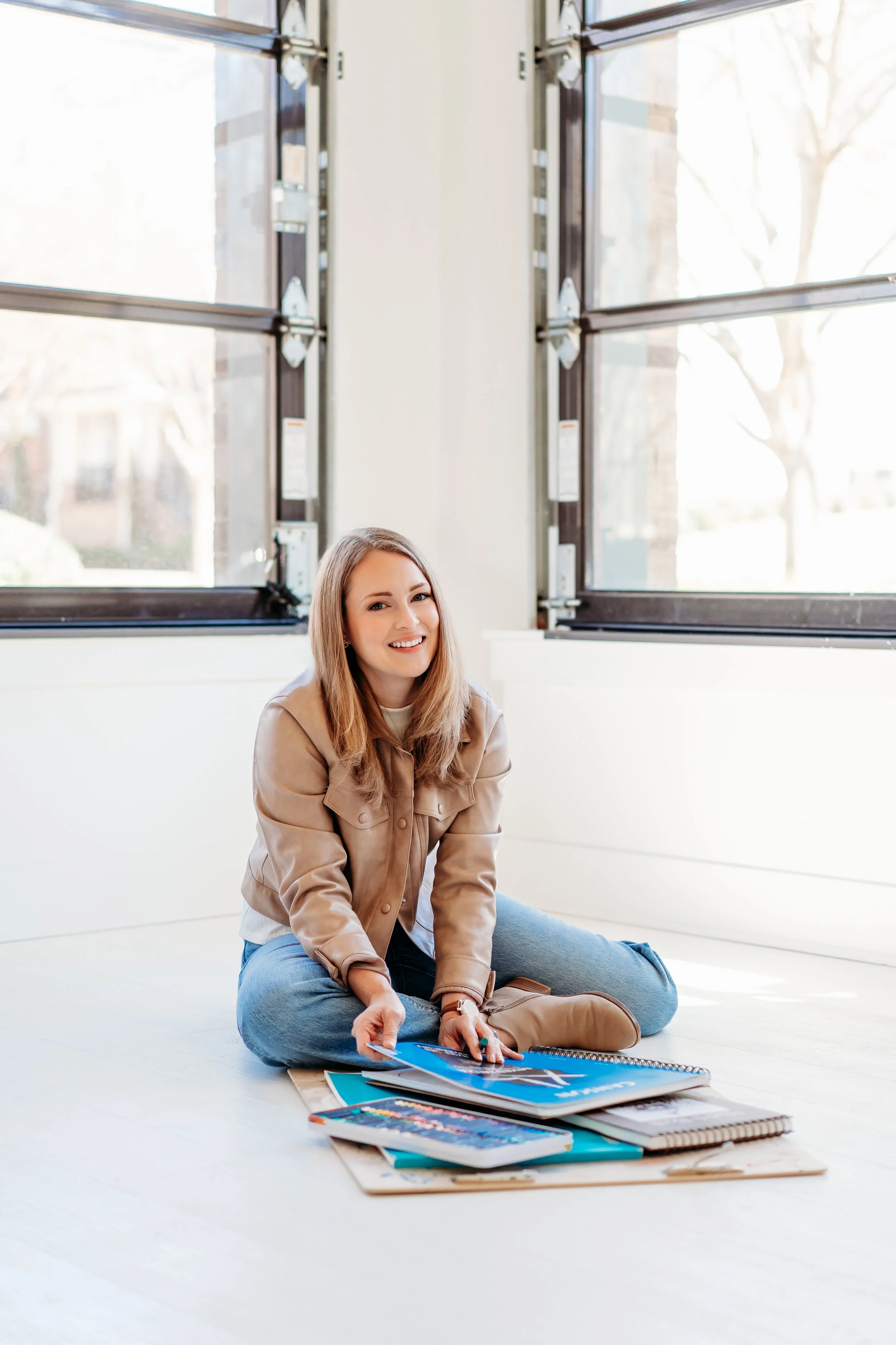 An art therapist in Marietta sitting cross-legged on the floor near large windows, smiling and looking at the camera. She has notebooks and art supplies spread out on the floor in front of her.